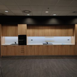 First Floor sp. z o.o - Nowoczesna zabudowa kuchenna z drewnianymi frontami, czarnym piekarnikiem w zabudowie i białym blatem, oświetlona od góry, widok na fragment ekspozycji w salonie meblowym.