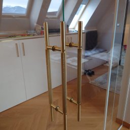 First Floor sp. z o.o - Szklane drzwi z dwoma złotymi uchwytami o teksturowanej powierzchni, widok na poddasze z oknami dachowymi i białe meble w tle.