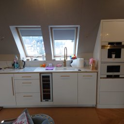 First Floor sp. z o.o - Biała zabudowa kuchenna z marmurowym blatem i złotymi detalami w jasnym wnętrzu pod skosem, z widokiem na miasto przez okna dachowe.