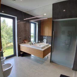 First Floor sp. z o.o - Nowoczesna łazienka w Krakowie z drewnianą szafką pod umywalką, dużym lustrem i szarą glazurą, widok na zieleń za oknem.