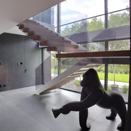 First Floor sp. z o.o - Nowoczesne schody wiszące z drewnianymi stopniami i szklaną balustradą w przestronnym holu z dużą figurą goryla