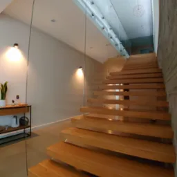Nowoczesne, drewniane schody wiszące z balustradą ze szkła, oświetlone lampami punktowymi, z widocznym fragmentem betonowej ściany i designerskimi lampami wiszącymi.