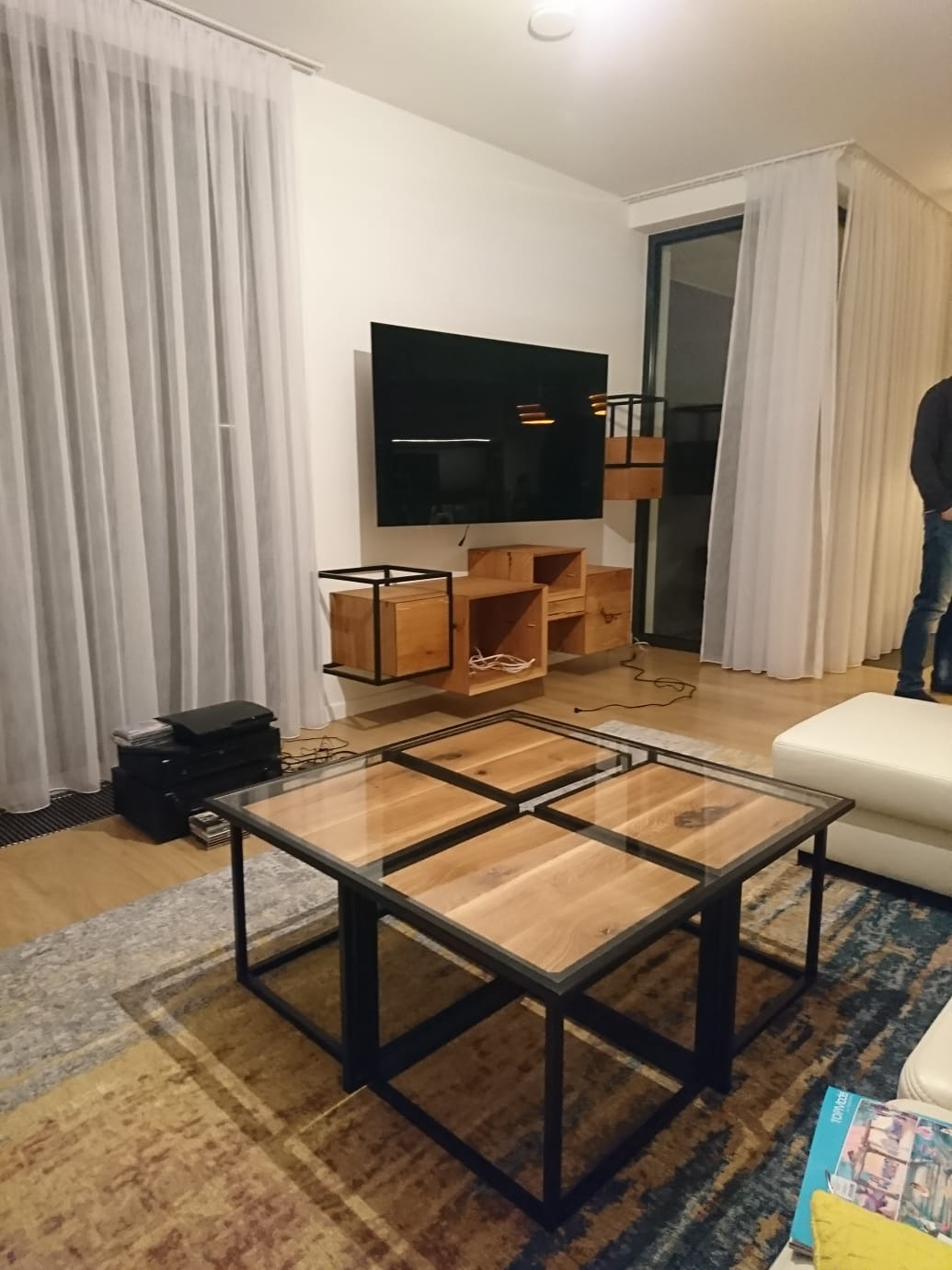 Nowoczesny salon z drewnianą szafką RTV wiszącą na ścianie, stolikiem kawowym z drewnianym blatem i czarną metalową ramą oraz długimi białymi zasłonami.