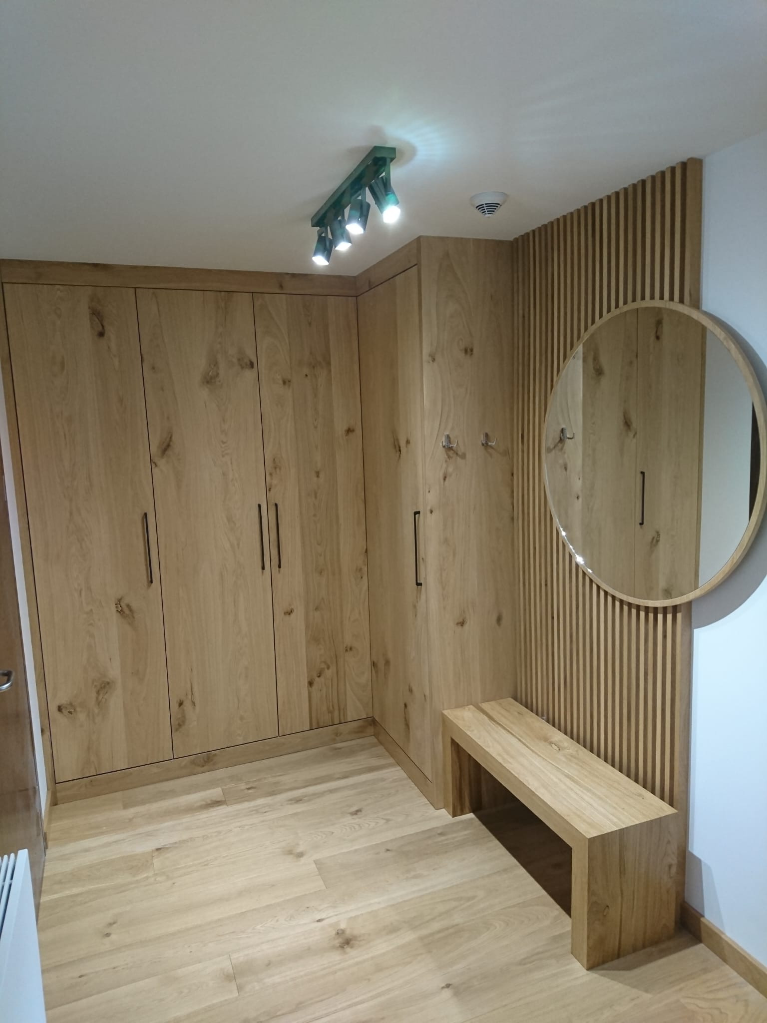 Jasna, drewniana szafa wnękowa z ciemnymi, pionowymi uchwytami, drewniana ławka i okrągłe lustro na ścianie z drewnianymi lamelami w przedpokoju.