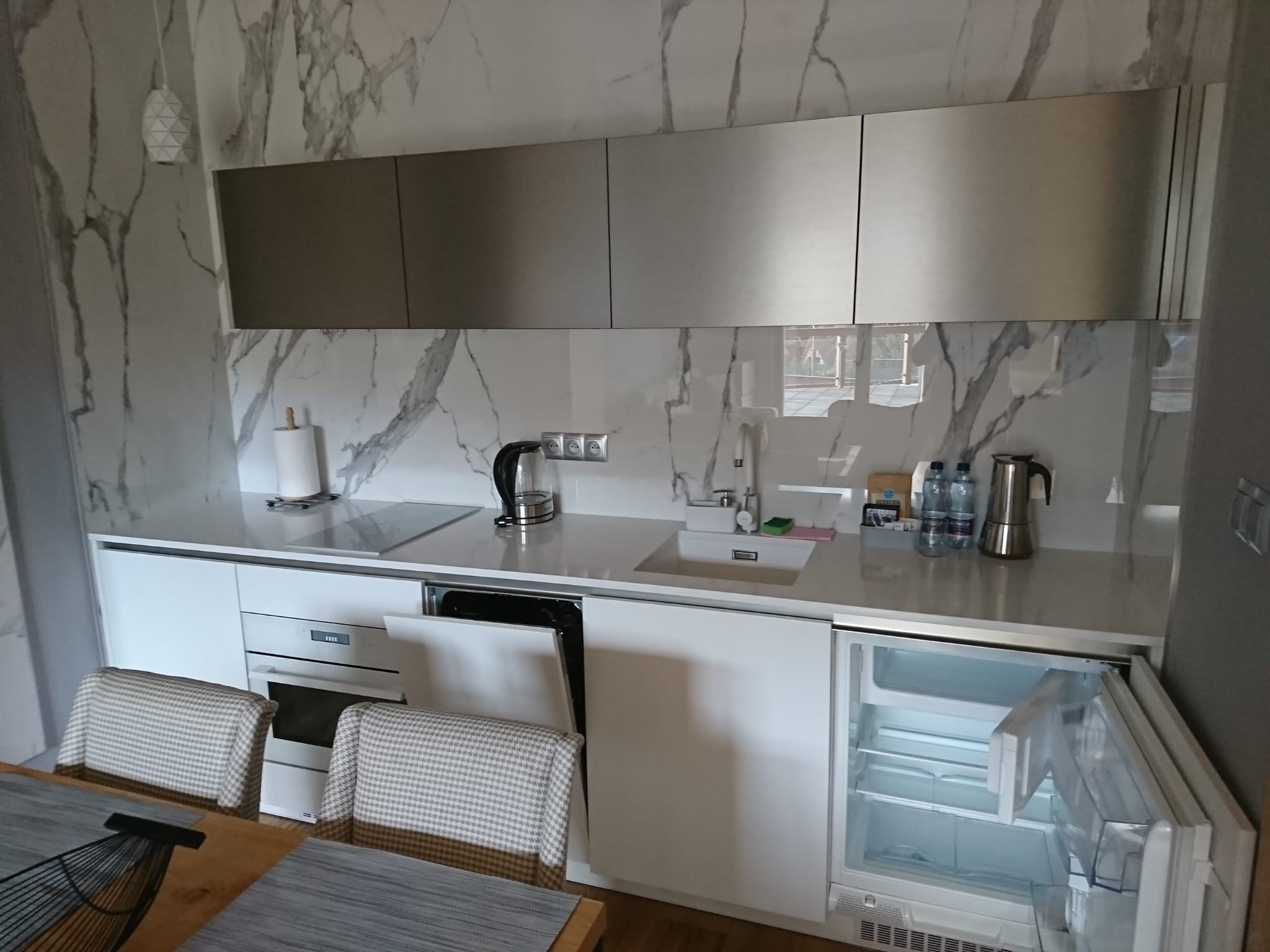 Nowoczesna kuchnia z białymi frontami i marmurowym backsplashiem, widoczny otwarty piekarnik i lodówka podblatowa, minimalistyczne oświetlenie.