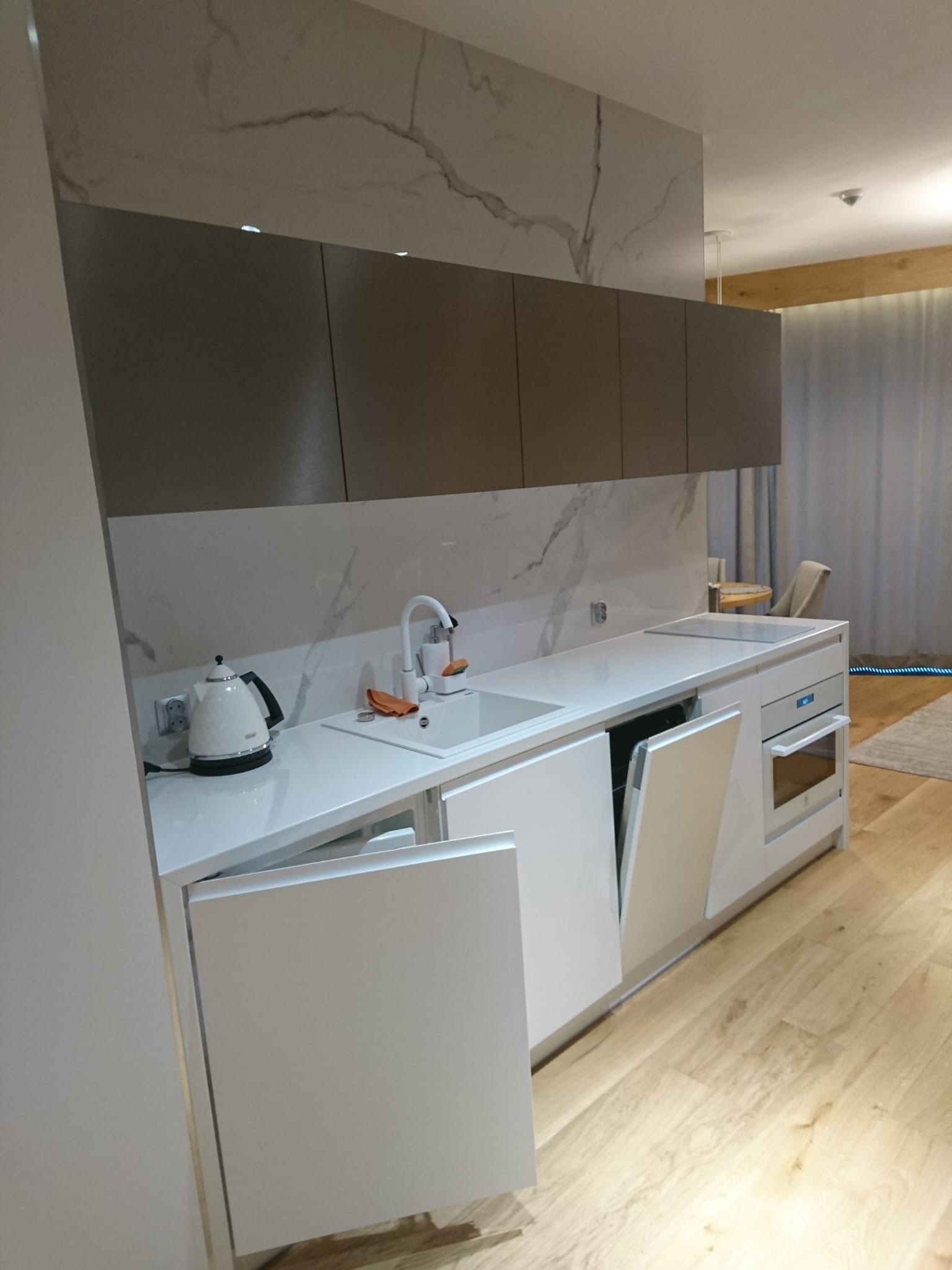 Nowoczesna kuchnia z białymi frontami i blatem z imitacją marmuru, otwartą zmywarką oraz drewnianą podłogą.