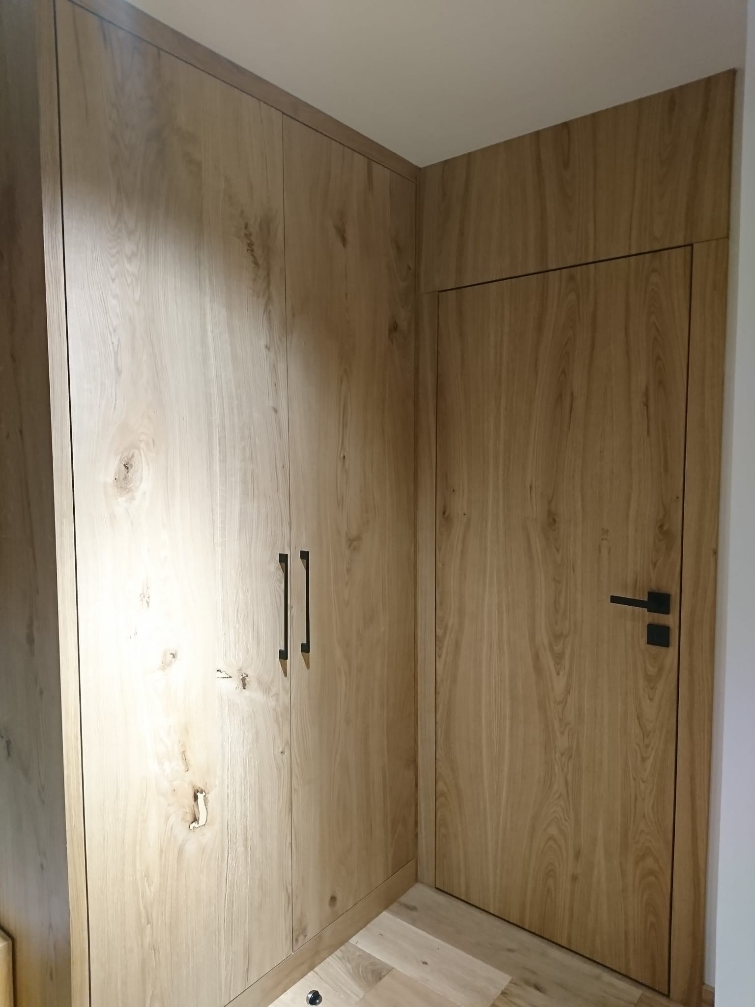 Zabudowa narożna z szafą i drzwiami o identycznym, jasnym wykończeniu drewnopodobnym, z czarnymi, minimalistycznymi uchwytami i klamką.