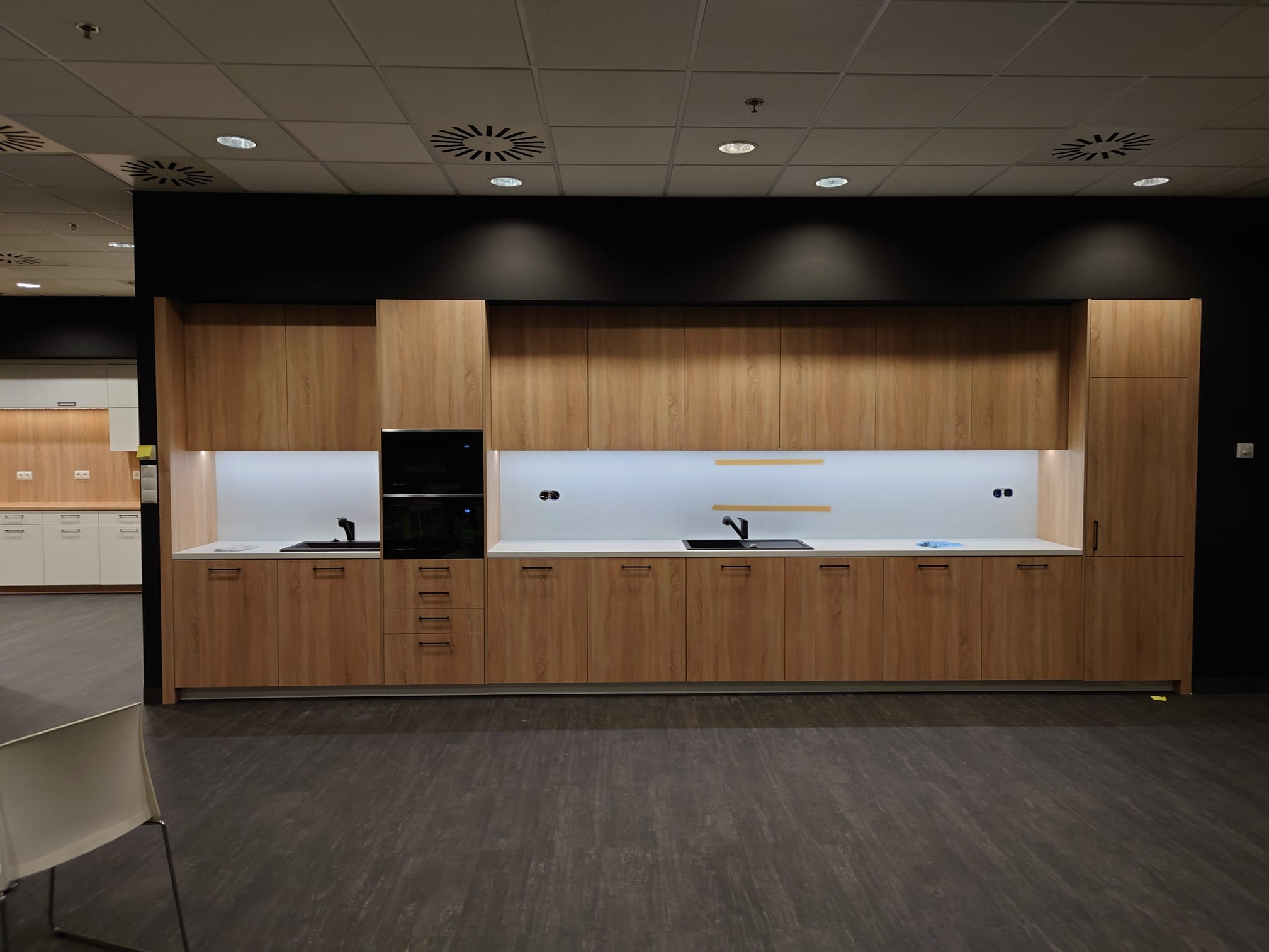 Nowoczesna zabudowa kuchenna z drewnianymi frontami, czarnym piekarnikiem w zabudowie i białym blatem, oświetlona od góry, widok na fragment ekspozycji w salonie meblowym.