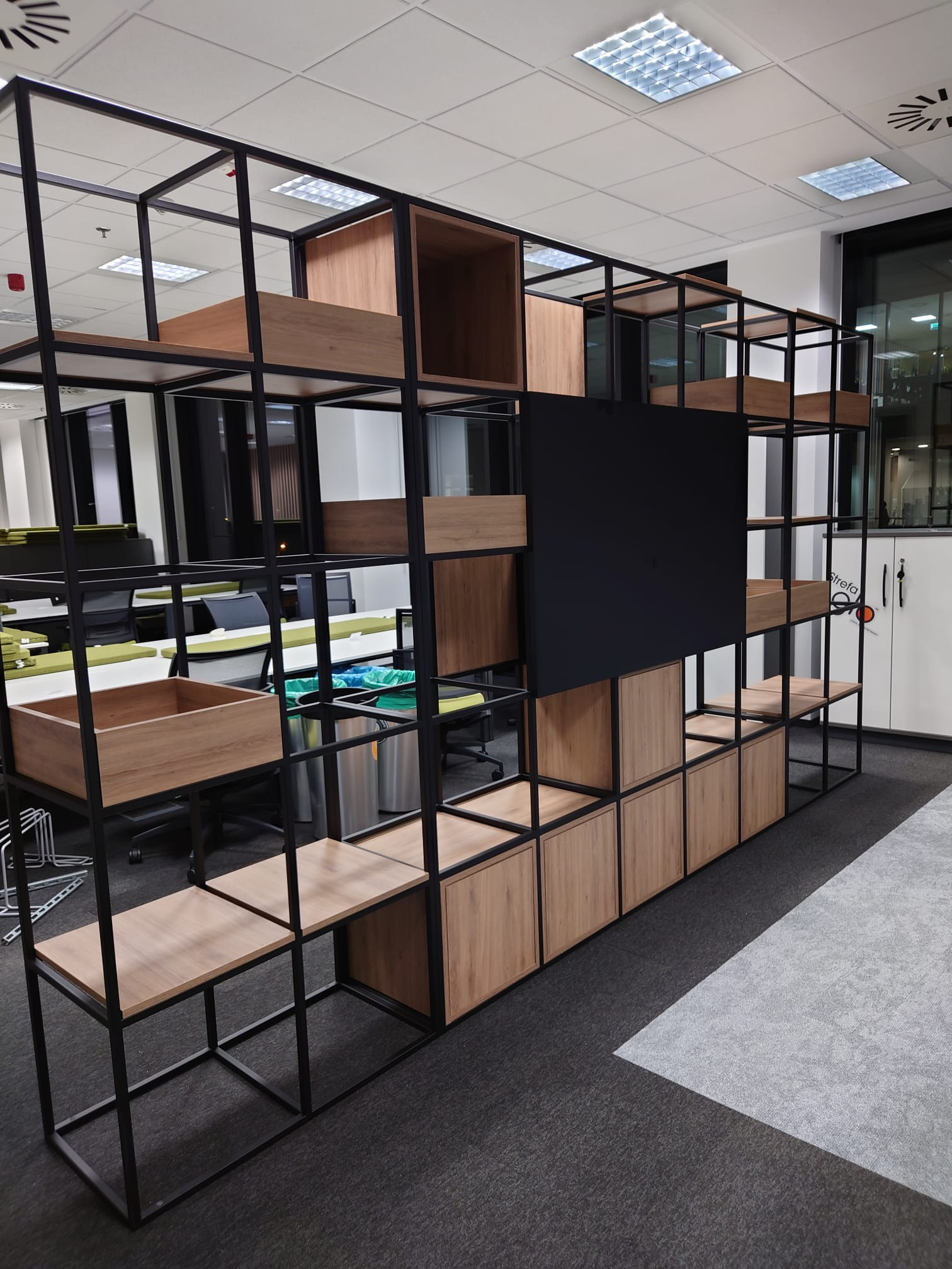 Regał biurowy z czarną metalową konstrukcją i drewnianymi elementami w odcieniach jasnego drewna, w tle widoczne biurka i krzesła biurowe.