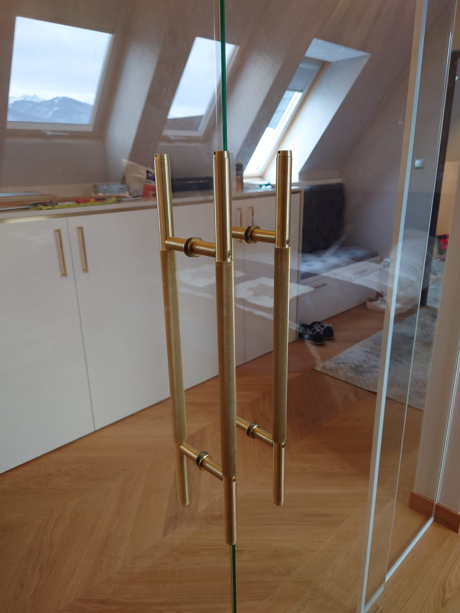 Szklane drzwi z dwoma złotymi uchwytami o teksturowanej powierzchni, widok na poddasze z oknami dachowymi i białe meble w tle.