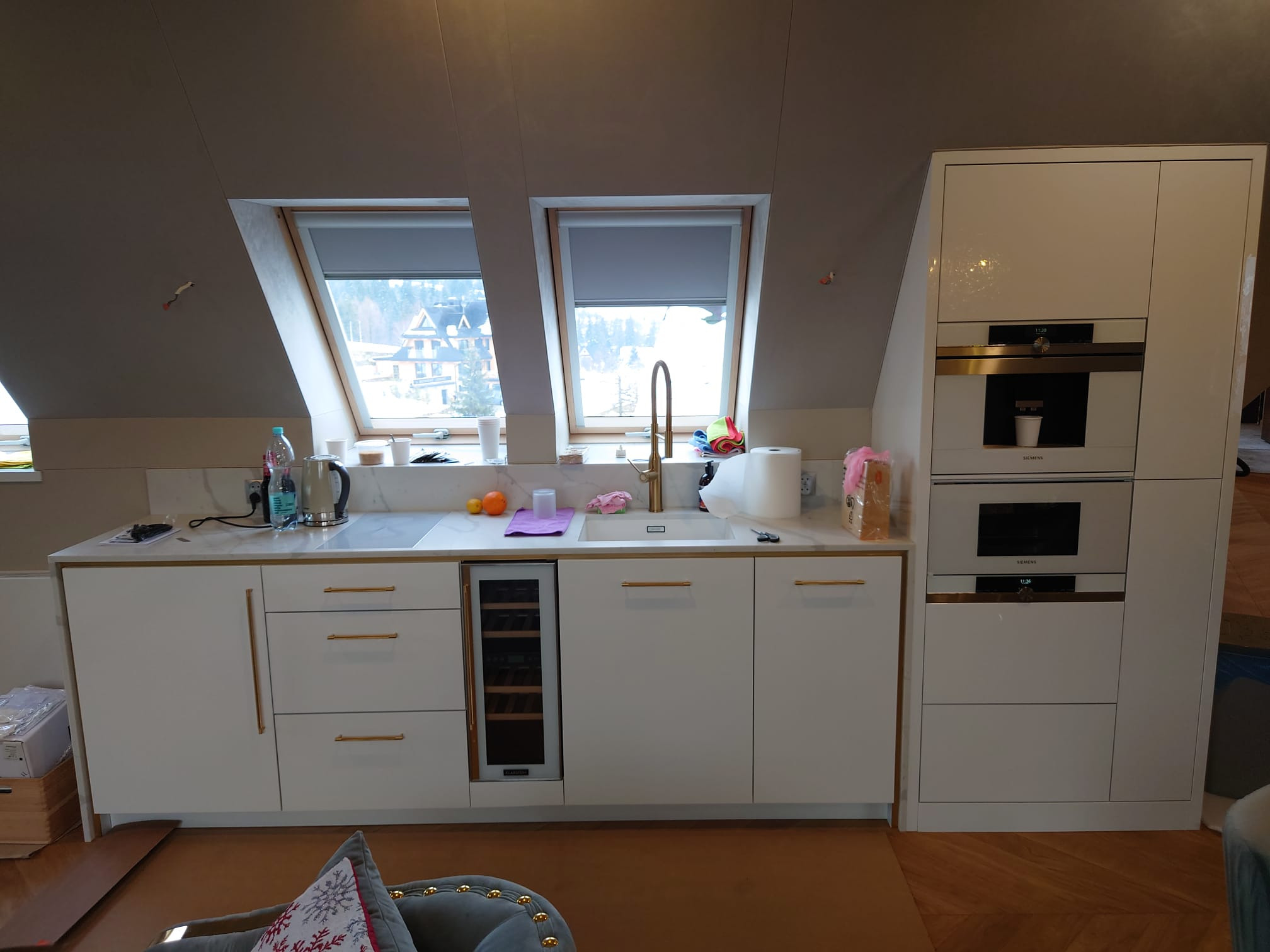 Biała zabudowa kuchenna z marmurowym blatem i złotymi detalami w jasnym wnętrzu pod skosem, z widokiem na miasto przez okna dachowe.