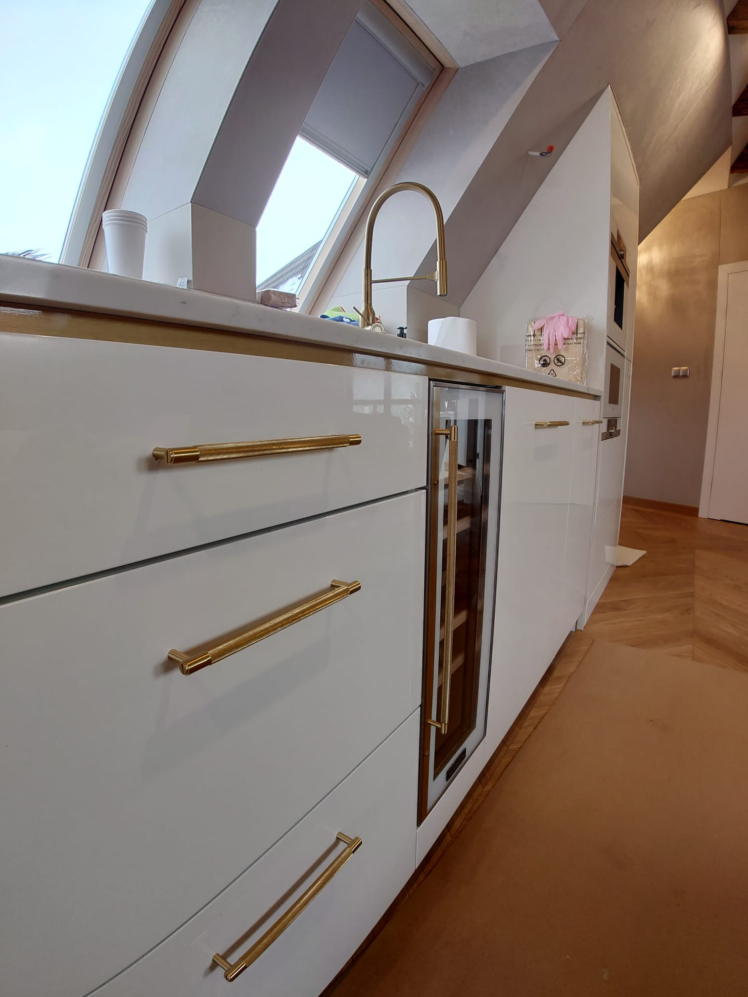 Nowoczesna kuchnia z białymi frontami szafek, złotymi uchwytami z radełkowanym wzorem, marmurowym blatem i wbudowaną chłodziarką do wina, umieszczona w pomieszczeniu ze skośnym oknem dachowym.