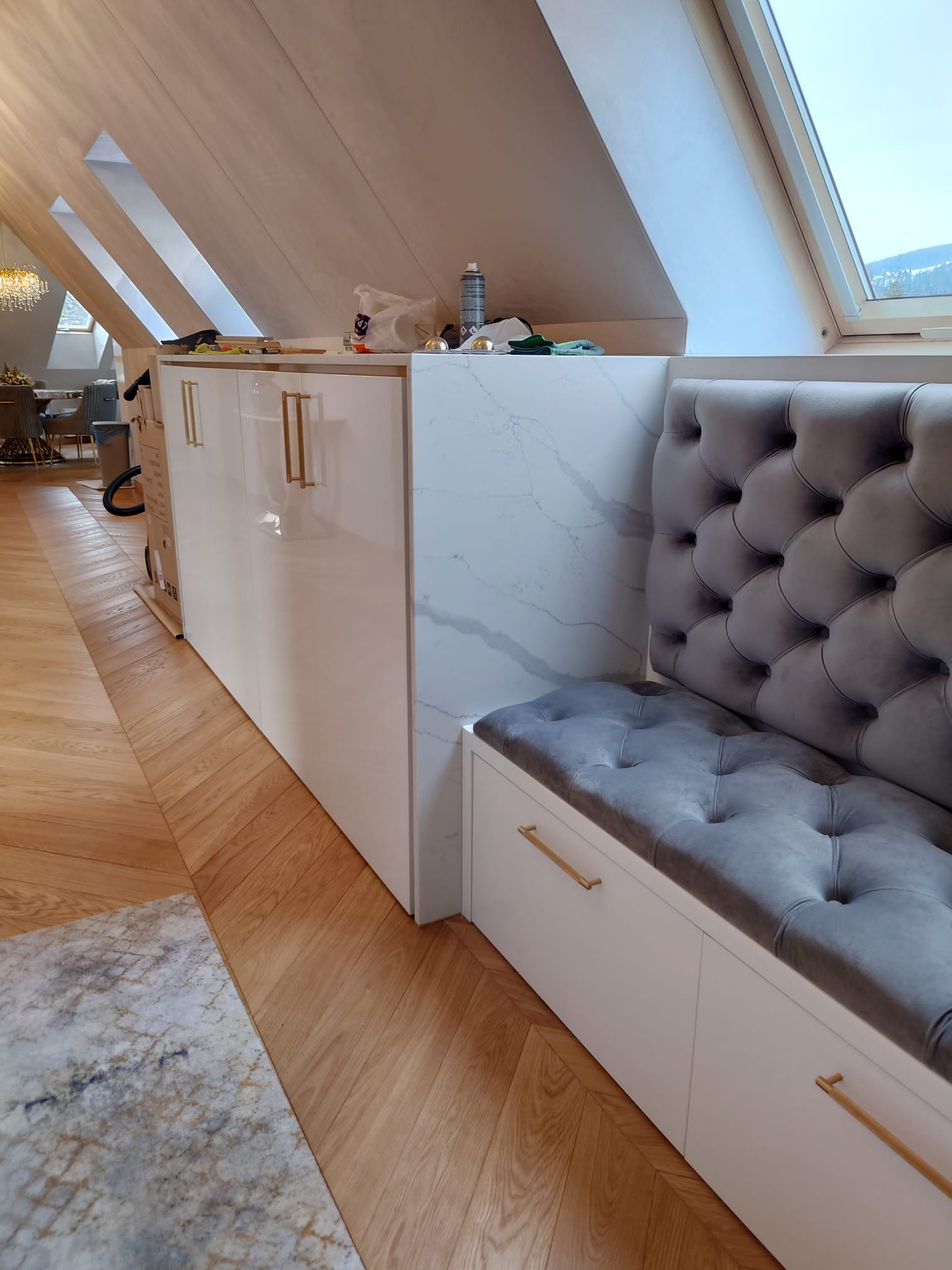 Biała zabudowa meblowa pod skosem dachu z szarym, pikowanym siedziskiem i złotymi uchwytami, widoczna część jadalni z kryształowym żyrandolem w tle.