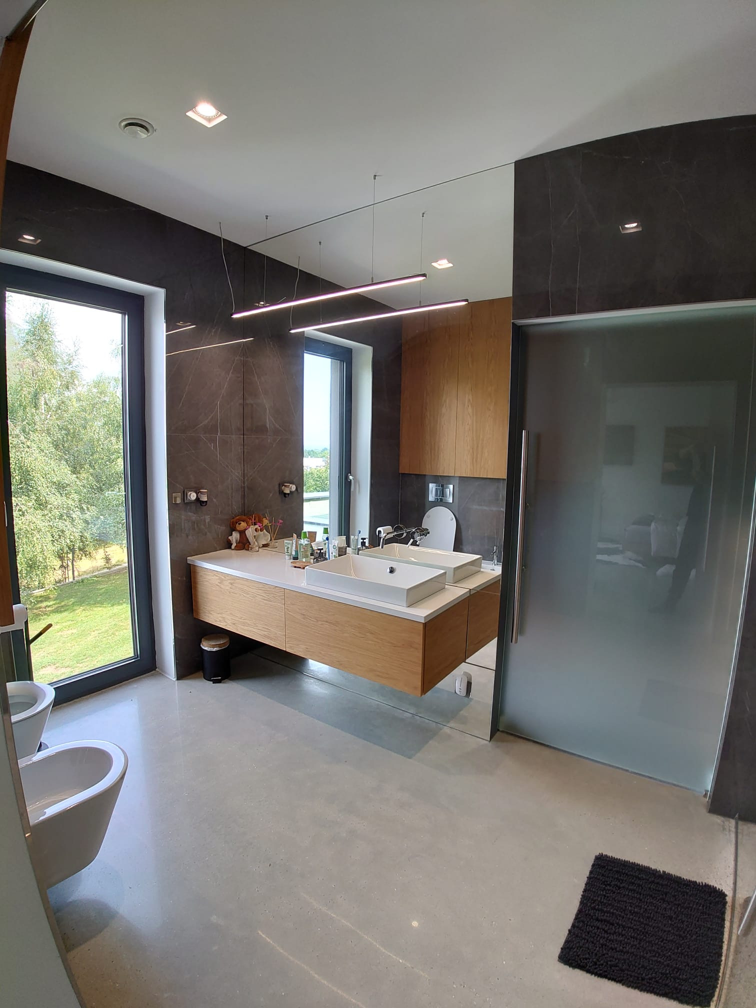 Nowoczesna łazienka w Krakowie z drewnianą szafką pod umywalką, dużym lustrem i szarą glazurą, widok na zieleń za oknem.