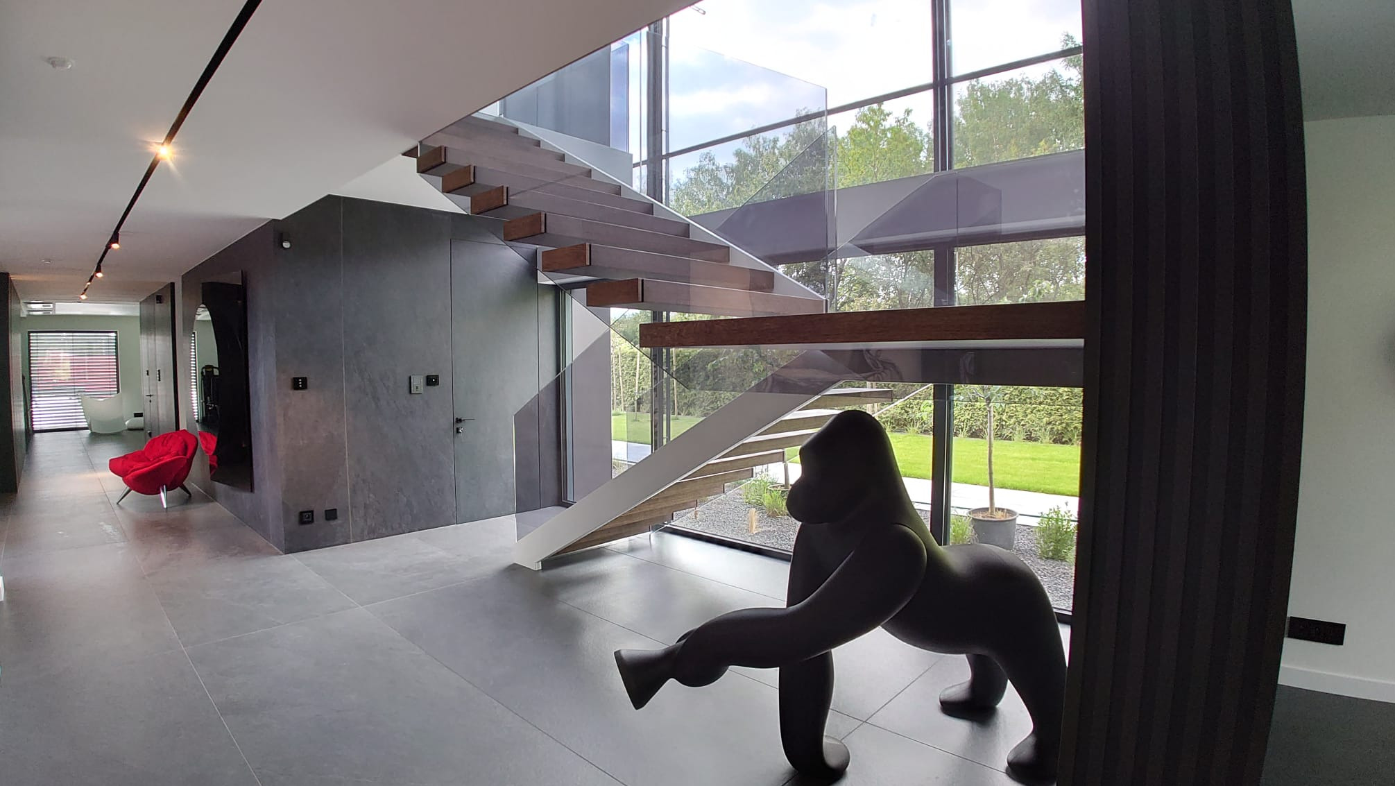 Nowoczesne schody wiszące z drewnianymi stopniami i szklaną balustradą w przestronnym holu z dużą figurą goryla