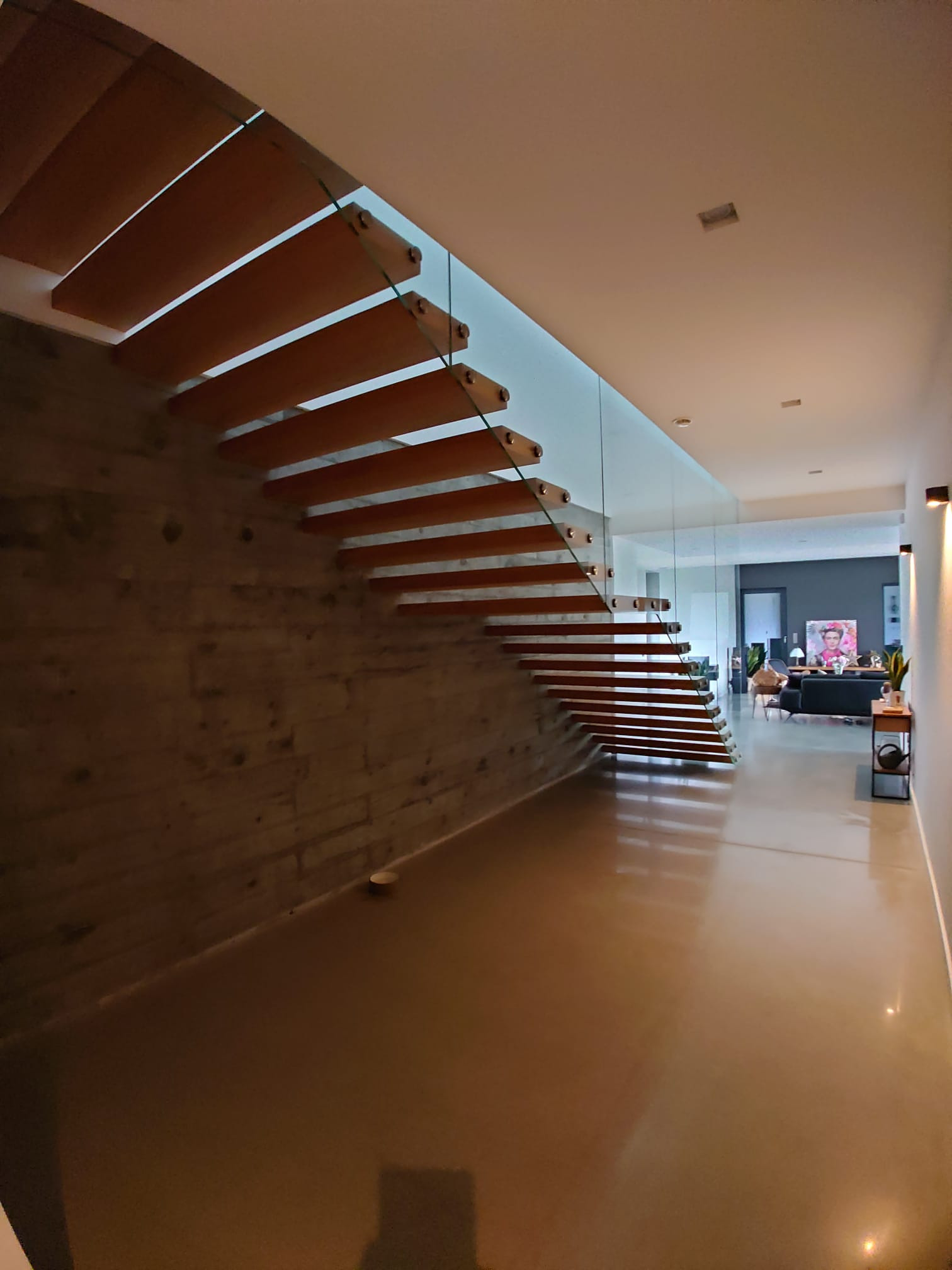 Nowoczesne schody drewniane wspornikowe z szklaną balustradą w minimalistycznym wnętrzu z betonową ścianą i polerowaną posadzką.