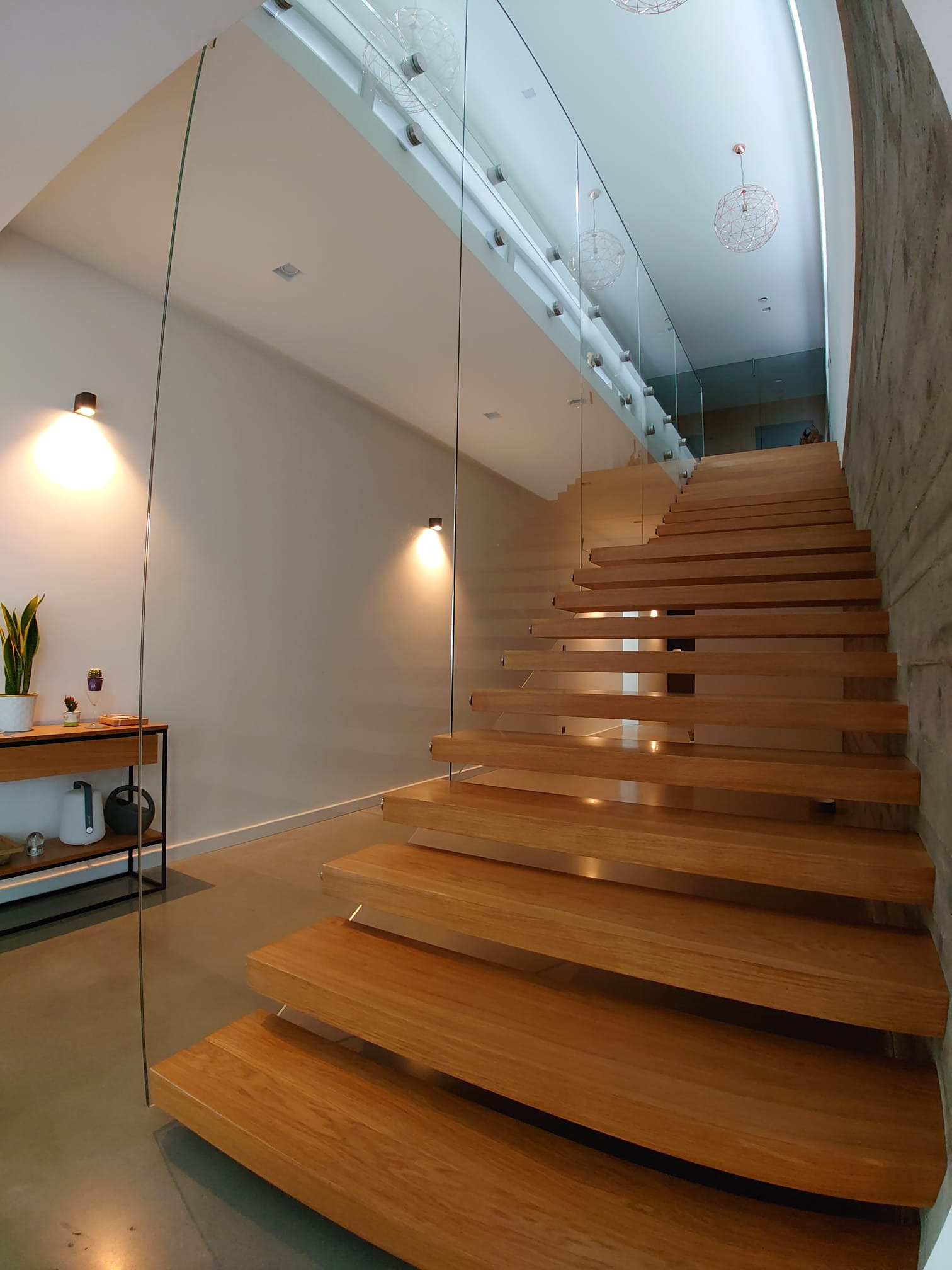 Nowoczesne, drewniane schody wiszące z balustradą ze szkła, oświetlone lampami punktowymi, z widocznym fragmentem betonowej ściany i designerskimi lampami wiszącymi.