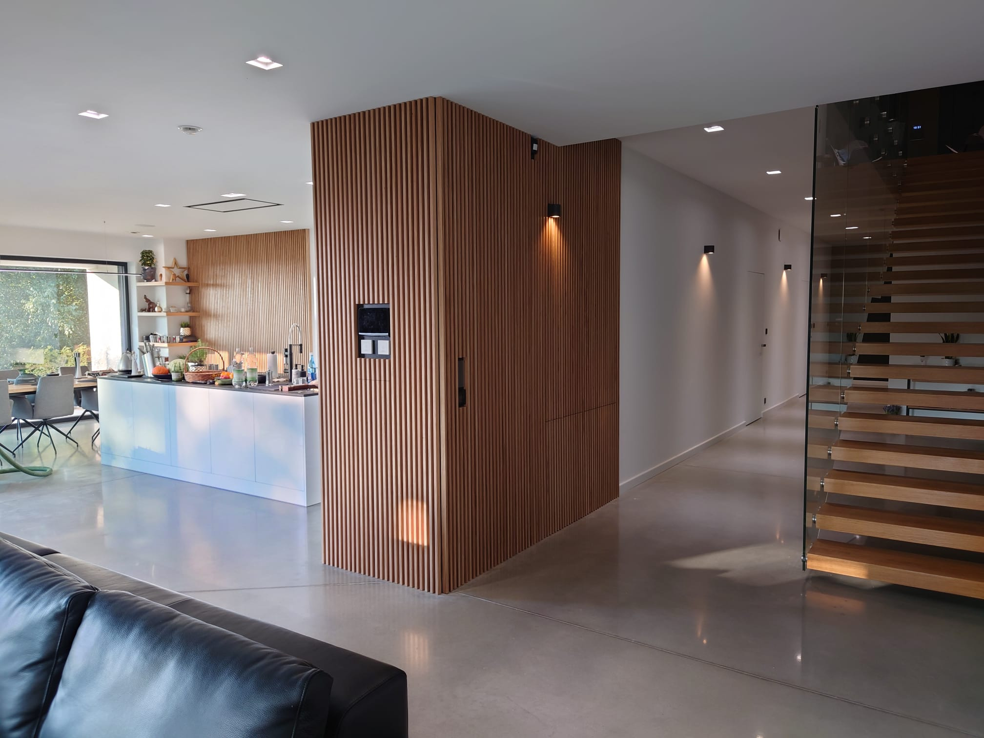 Wnętrze domu z nowoczesną kuchnią z białą wyspą, drewnianą ścianą z pionowych listew, widok na szklane schody i minimalistyczny korytarz z oświetleniem punktowym.