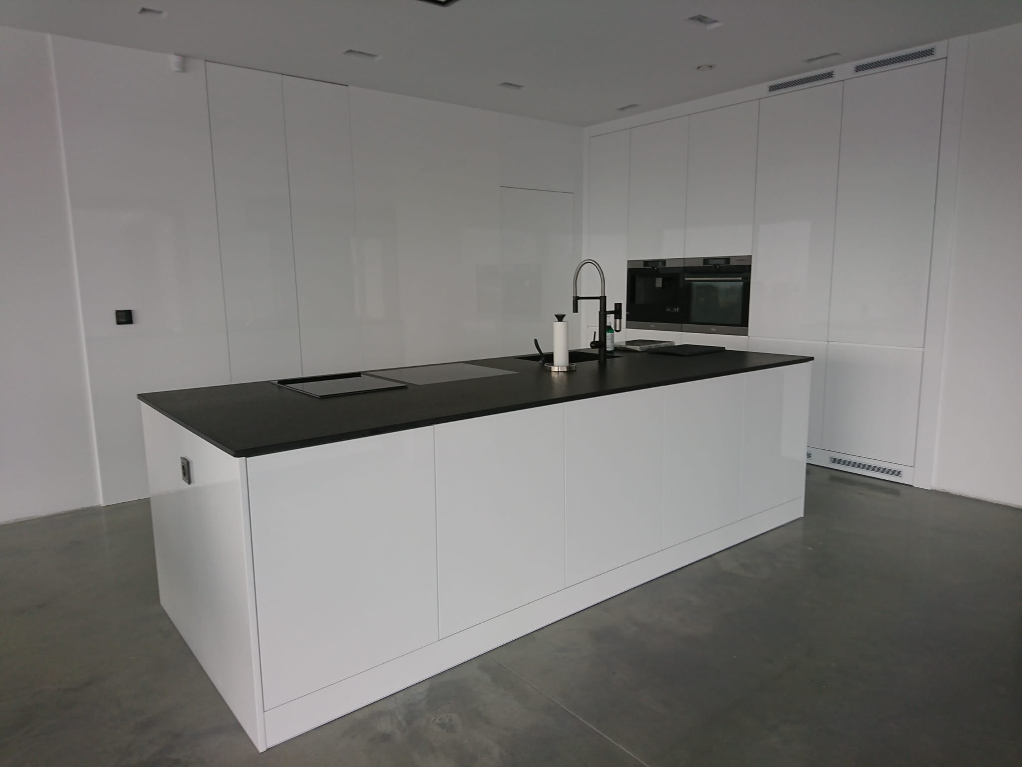 Minimalistyczna kuchnia z białymi, lakierowanymi frontami i czarnym blatem, wyspa kuchenna z wbudowaną płytą indukcyjną i zlewozmywakiem, zabudowa sięgająca sufitu z ukrytymi szafkami i piekarnikiem.