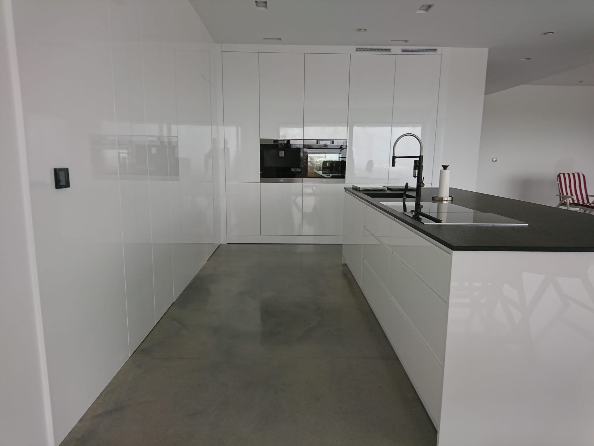 Minimalistyczna kuchnia z białymi, lakierowanymi frontami, czarnym blatem i szarą posadzką, widok na zabudowane sprzęty AGD.