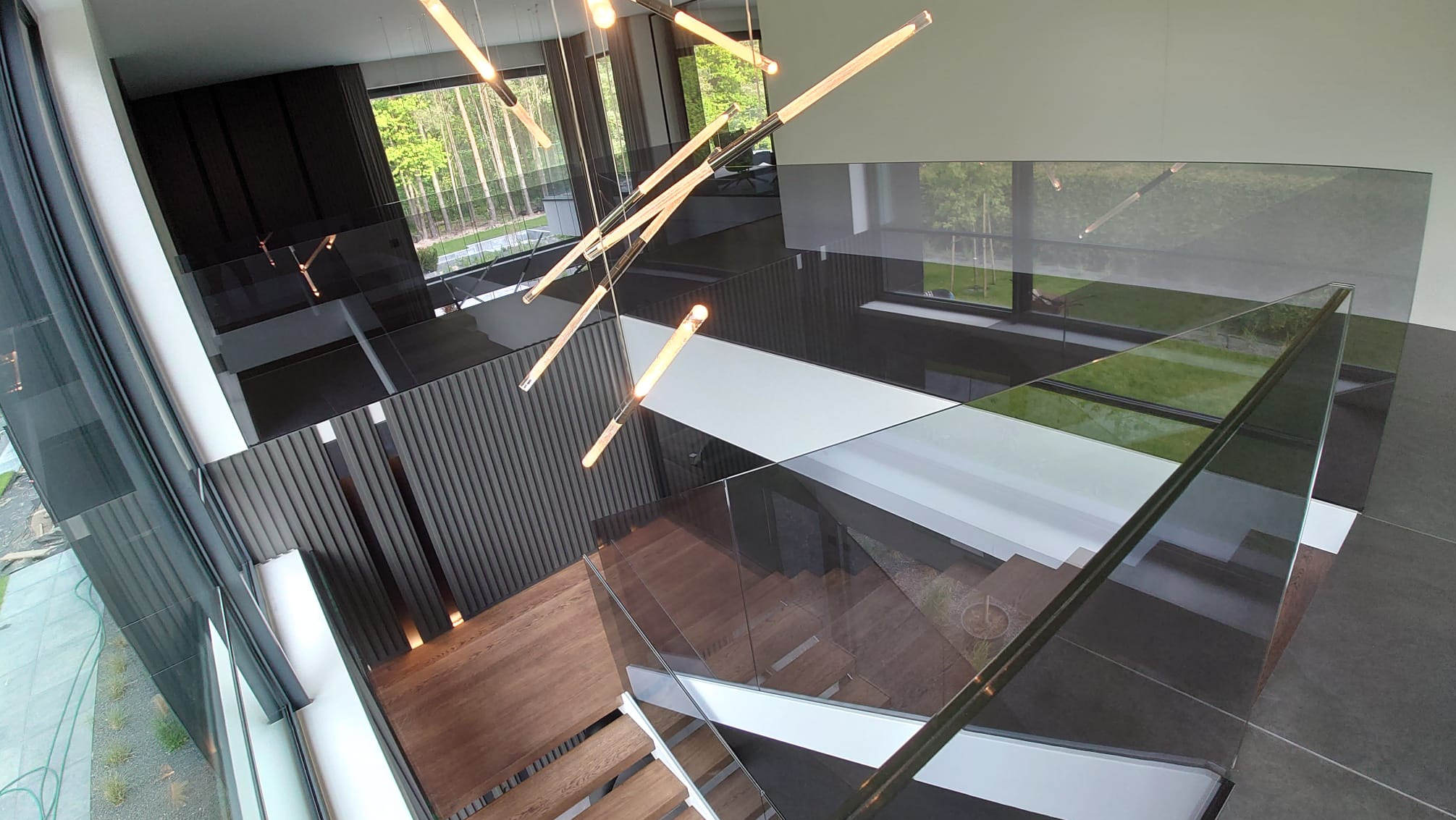 Nowoczesne schody drewniane z przeszklonymi balustradami, widok z góry, designerska lampa wisząca z podłużnymi żarówkami, duże okna z widokiem na zieleń.