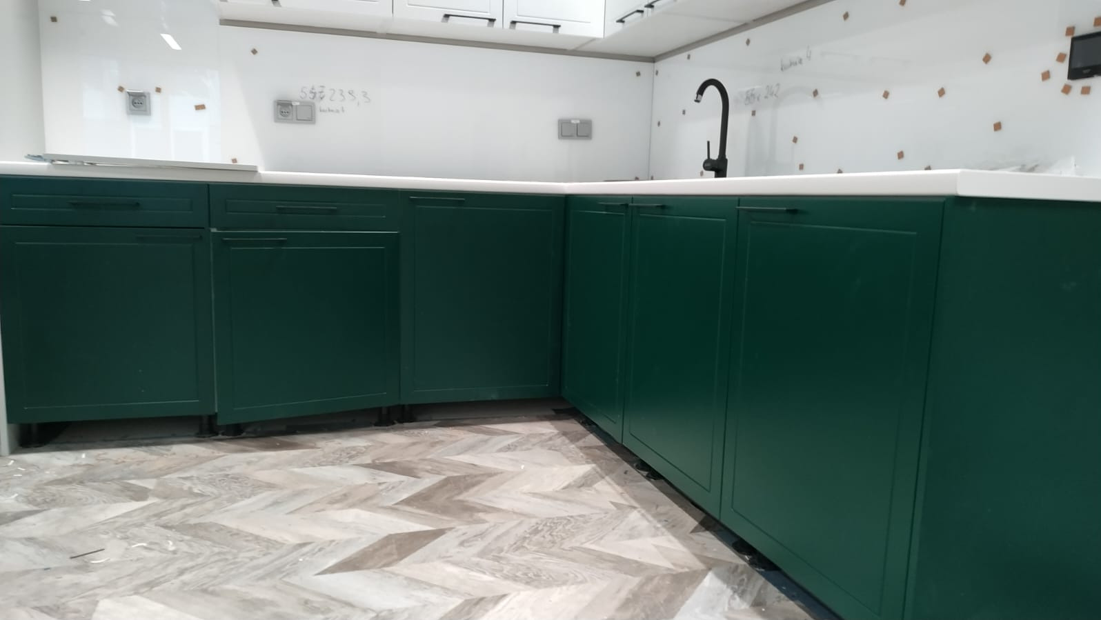 Fragment zielonej zabudowy kuchennej z białym blatem i czarną armaturą, widoczne ślady po remoncie na ścianie i podłodze.
