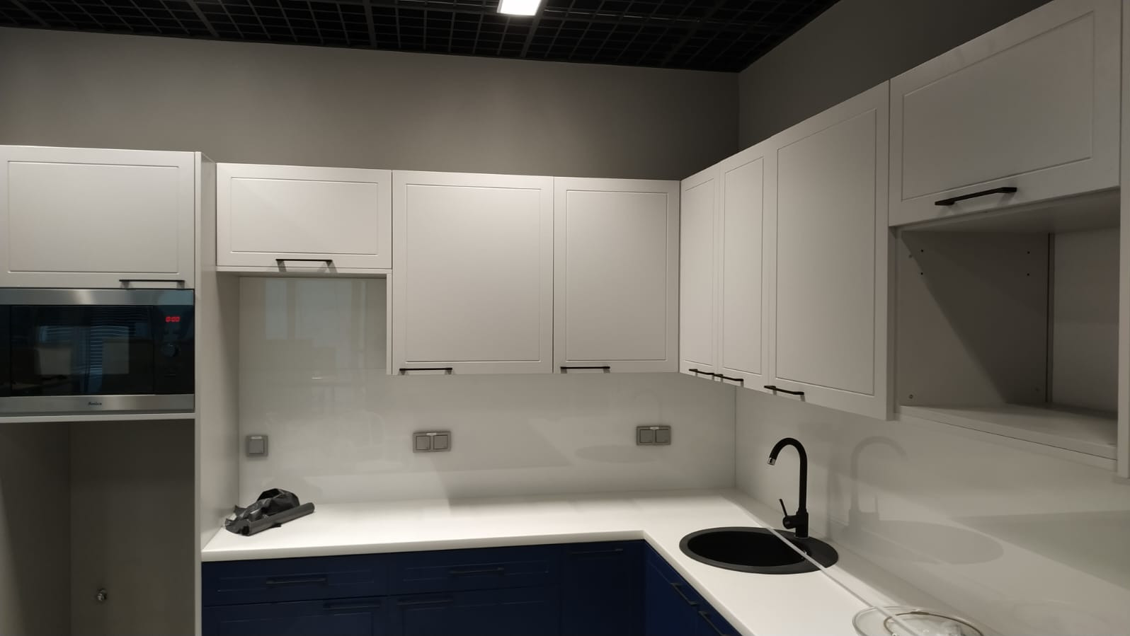 Nowoczesna kuchnia z białymi szafkami górnymi z czarnymi uchwytami, granatowymi szafkami dolnymi, białym blatem i czarnym zlewem oraz wbudowaną mikrofalówką.