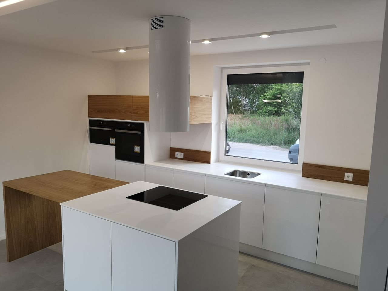 Nowoczesna kuchnia z białą wyspą, drewnianymi akcentami na szafkach i blacie, cylindrycznym okapem i dużym oknem z widokiem na zieleń.