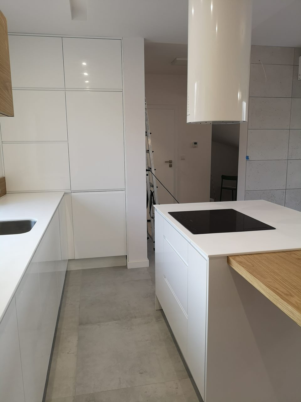 Minimalistyczna, biała kuchnia z wyspą i drewnianym blatem, widok z niskiej perspektywy, drabina w tle sugeruje etap montażu.