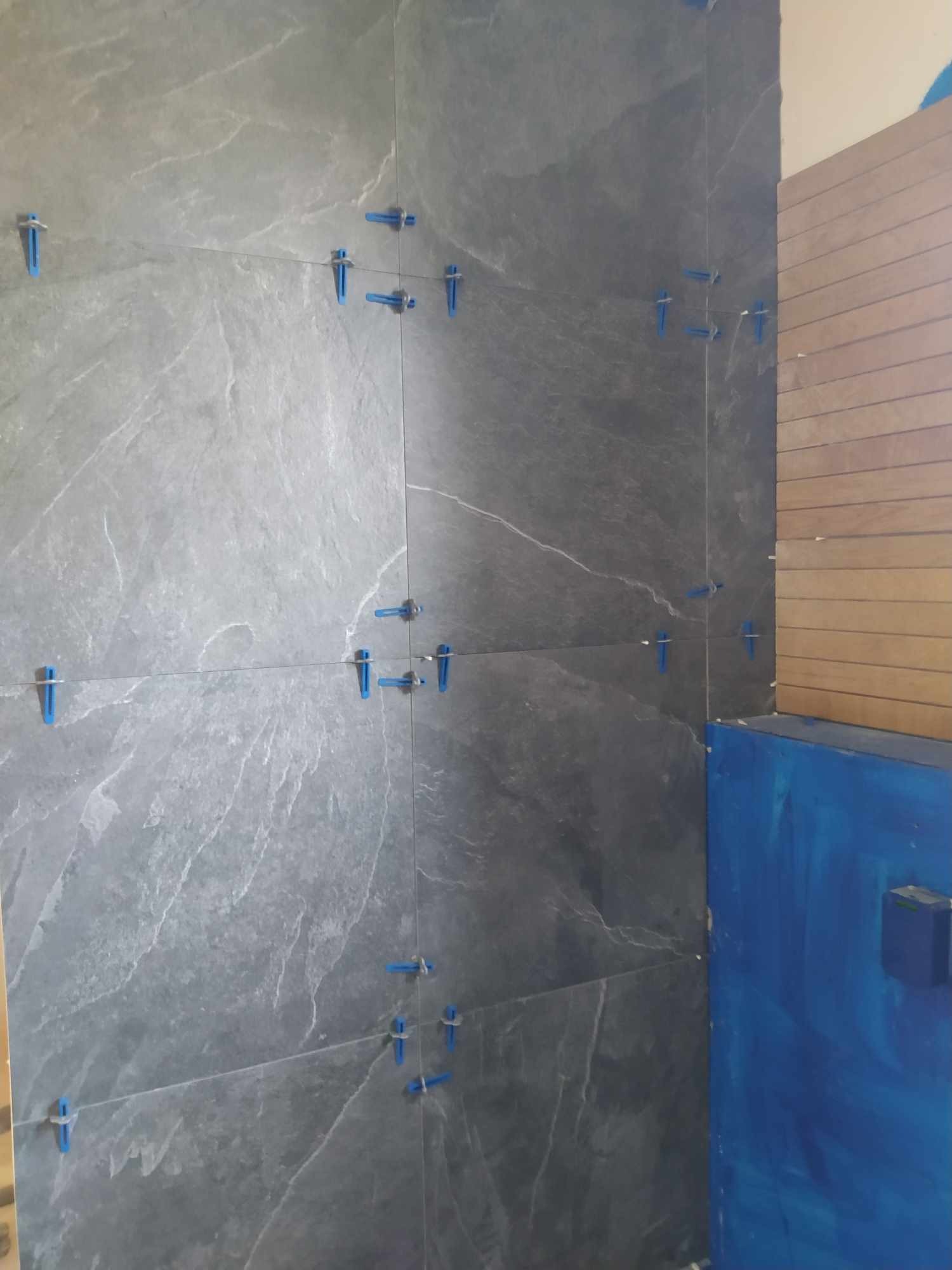 Szare płytki imitujące kamień na ścianie, ułożone z użyciem niebieskich krzyżyków dystansowych i poziomicy laserowej, widoczna zabezpieczona niebieską folią część ściany i fragment drewnianej...