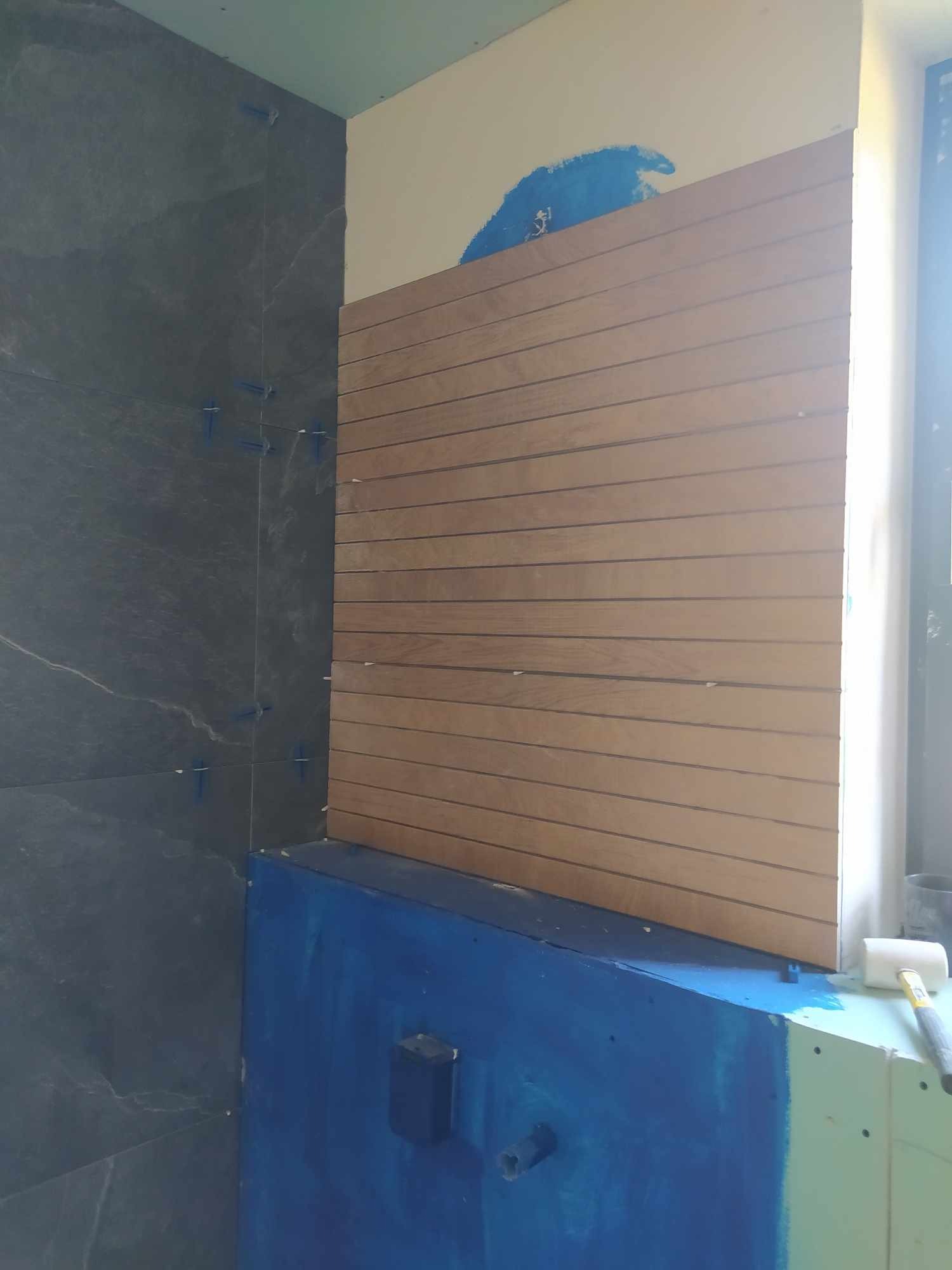 Fragment łazienki w trakcie prac: ciemne płytki imitujące kamień z krzyżykami dystansowymi, drewnopodobne panele na ścianie, niebieska hydroizolacja na wannie, gumowy młotek obok