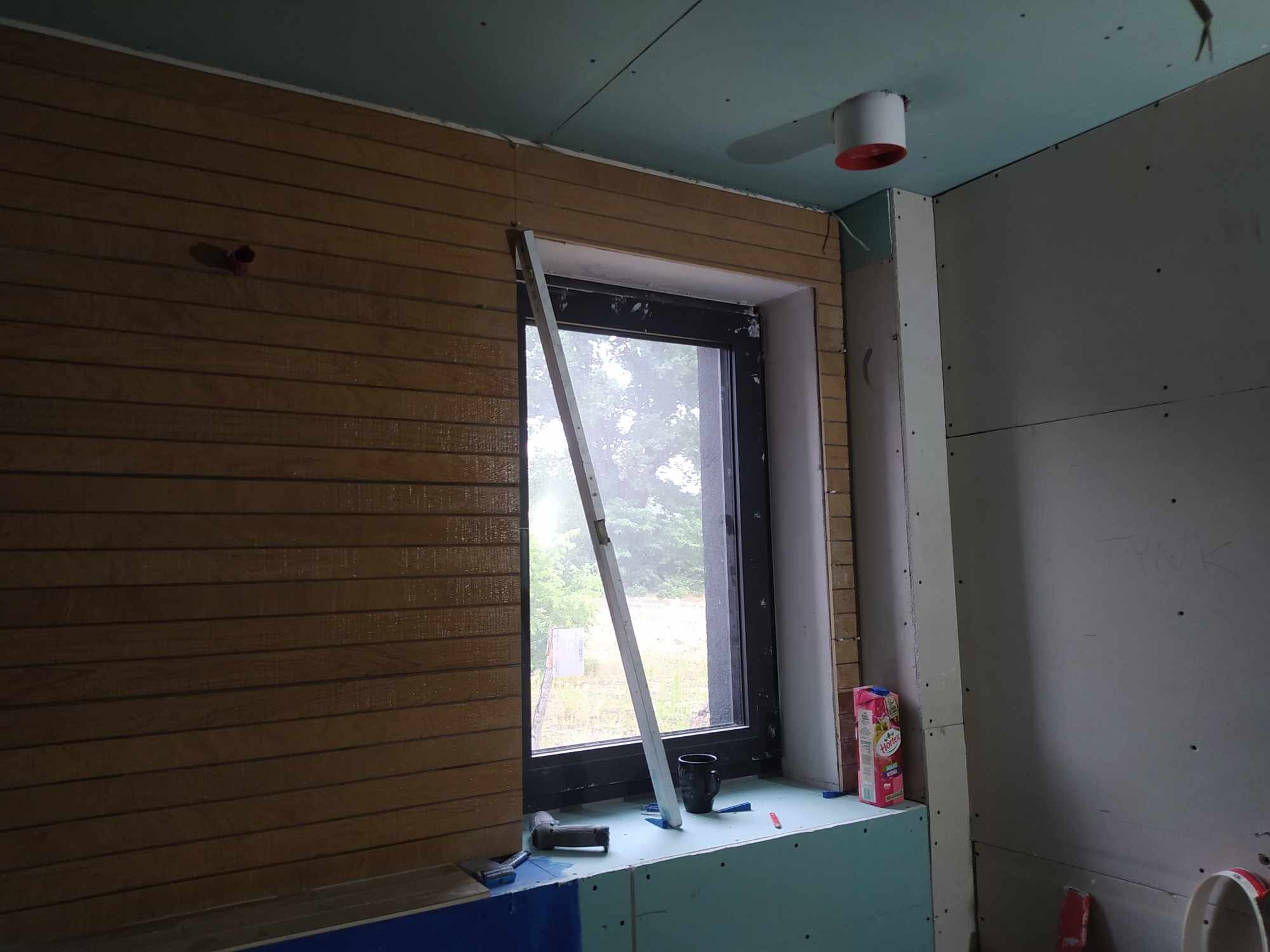 Łazienka w trakcie prac: montaż drewnianej boazerii przy oknie, ściany wykończone płytami gipsowo-kartonowymi, widoczny poziomica oparta o ramę okna, narzędzia i karton soku na parapecie.