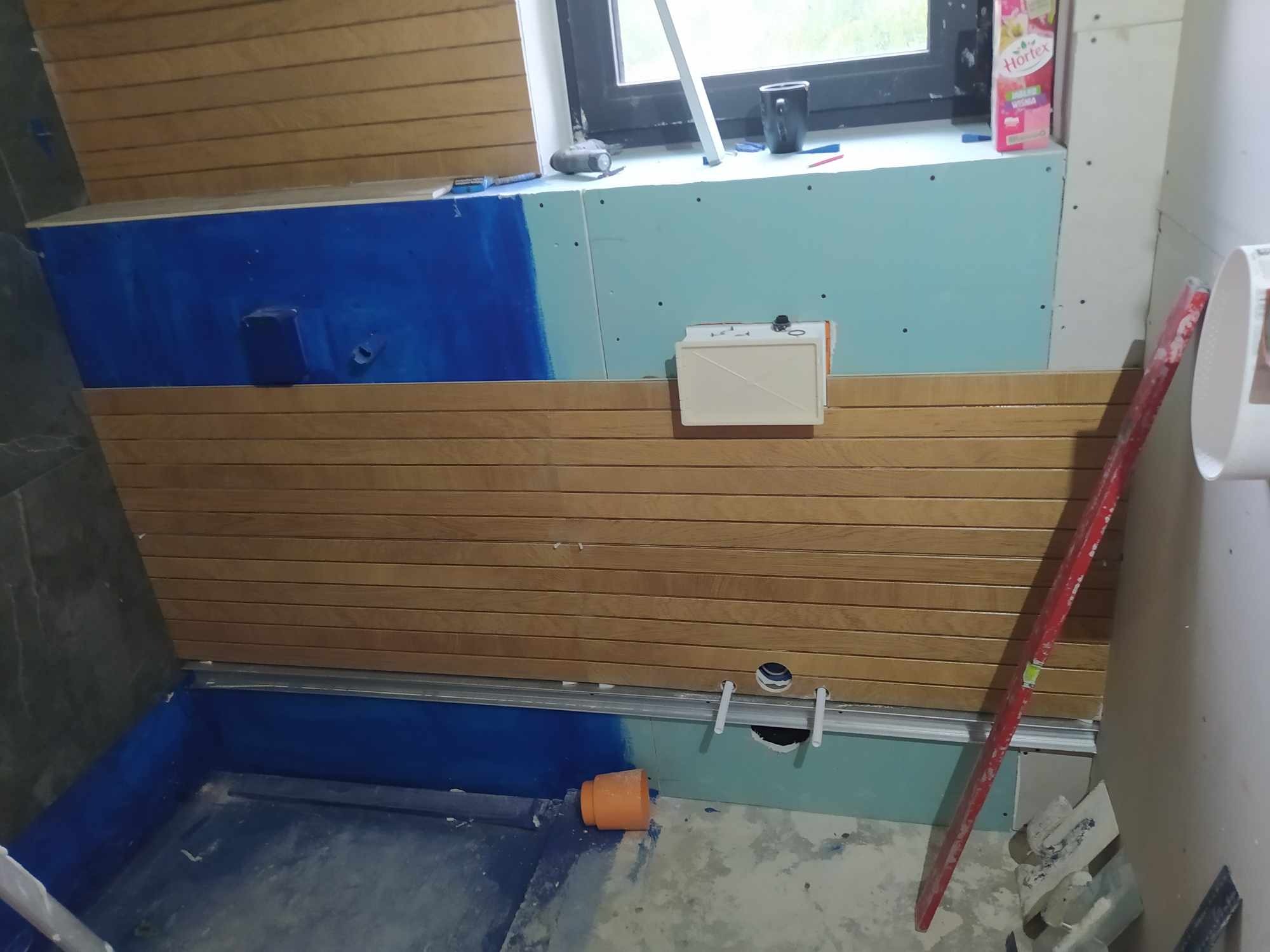 Łazienka w trakcie remontu: hydroizolacja brodzika w kolorze niebieskim, częściowo obłożona panelami drewnopodobnymi, widoczne płyty karton-gips, poziomica i narzędzia, butelka soku Hortex...