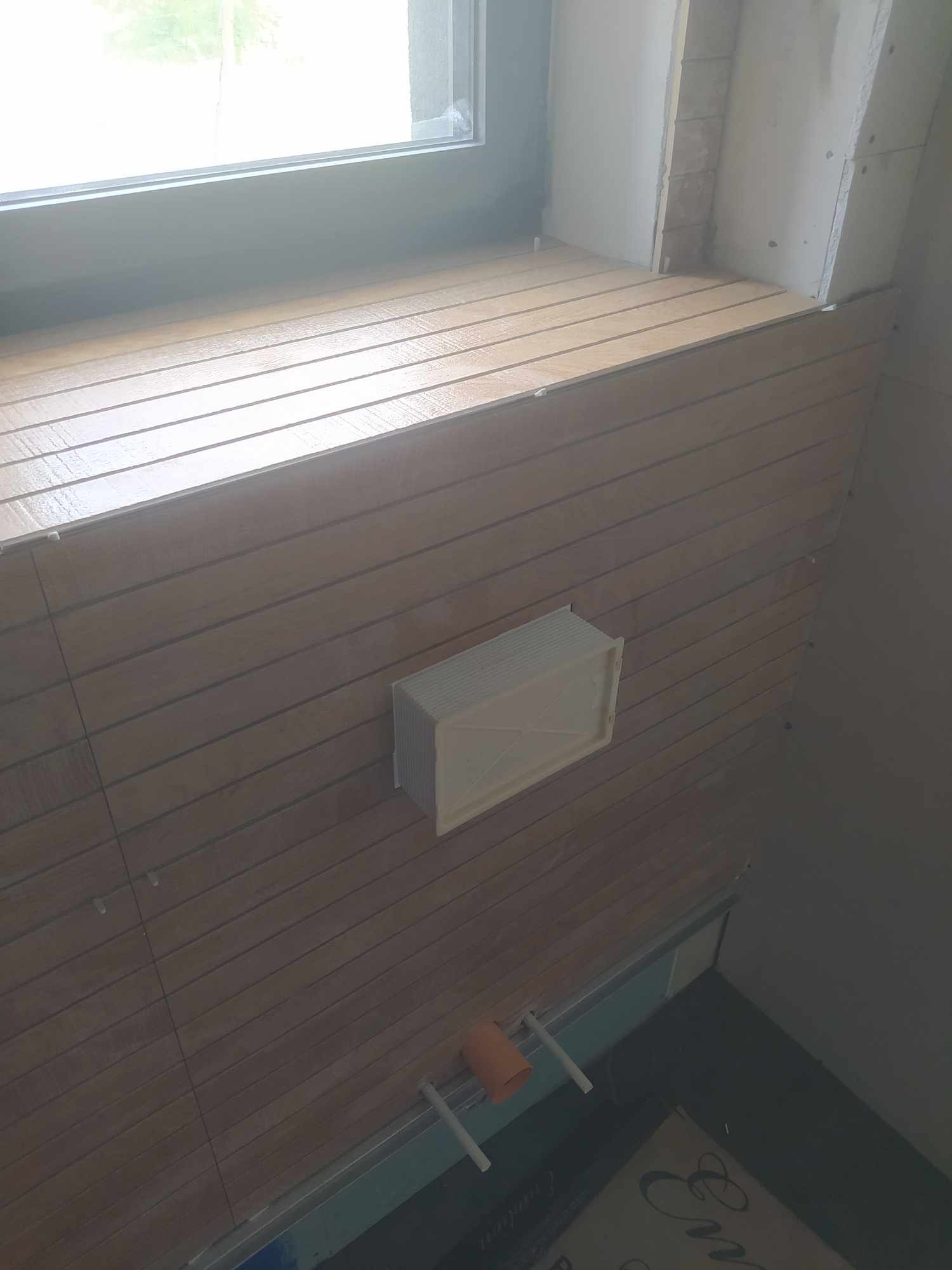 Ściana łazienki wyłożona poziomymi panelami imitującymi drewno, z zamontowanym anemostatem wentylacyjnym i widocznymi elementami instalacji hydraulicznej u dołu ściany, pod oknem.