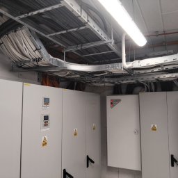 Torway sp. z o.o. - Rozbudowana instalacja elektryczna w budynku: kable w korytach, szafy sterownicze z oznaczeniami ostrzegawczymi. Widoczne elementy oświetlenia i okablowania.