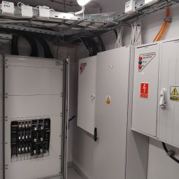 Torway sp. z o.o. - Rozdzielnia elektryczna z otwartymi drzwiami, widoczne zabezpieczenia i okablowanie. Tablice ostrzegawcze i oznaczenia. Instalacja w pomieszczeniu technicznym.