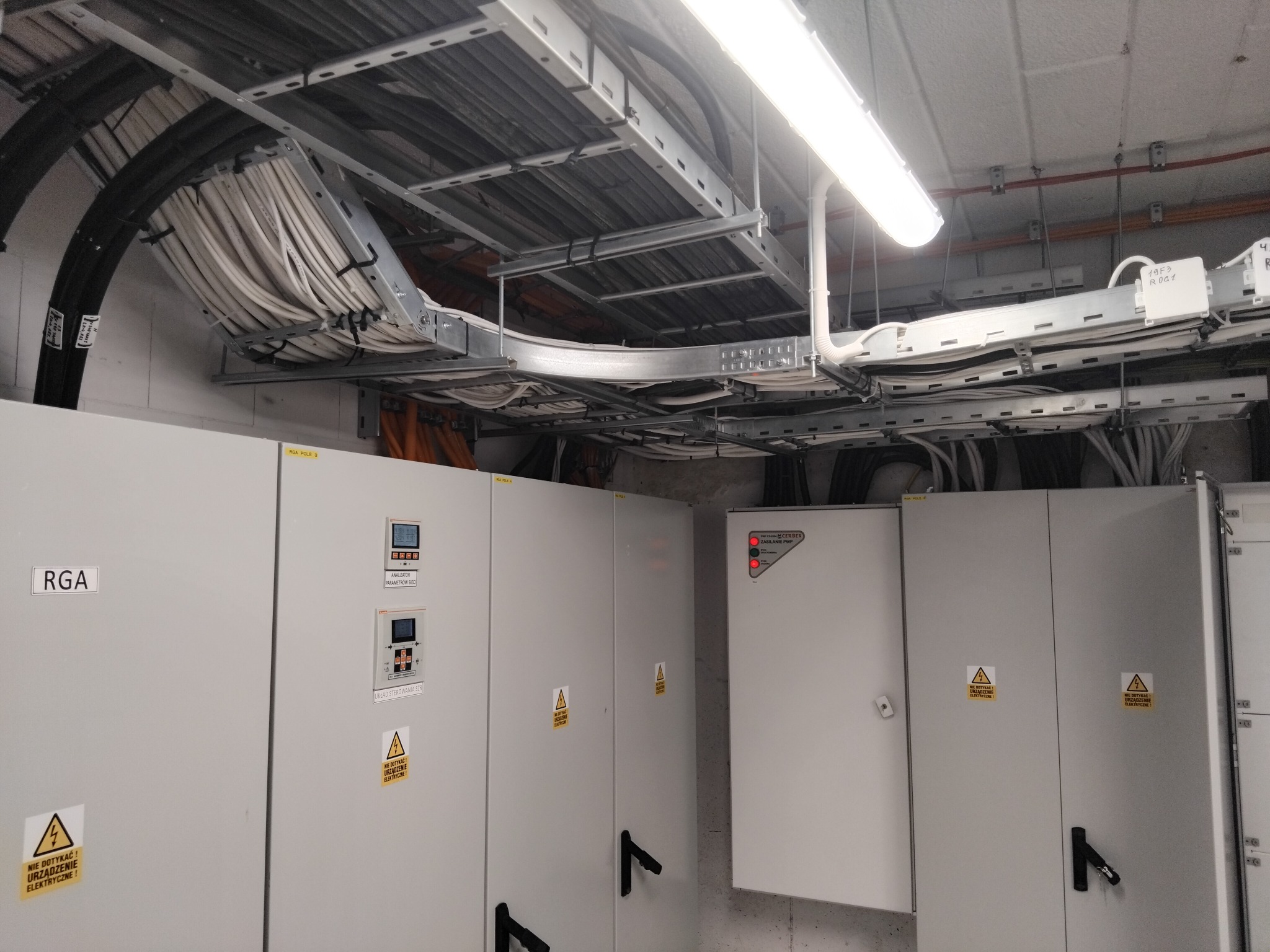 Rozbudowana instalacja elektryczna w budynku: kable w korytach, szafy sterownicze z oznaczeniami ostrzegawczymi. Widoczne elementy oświetlenia i okablowania.