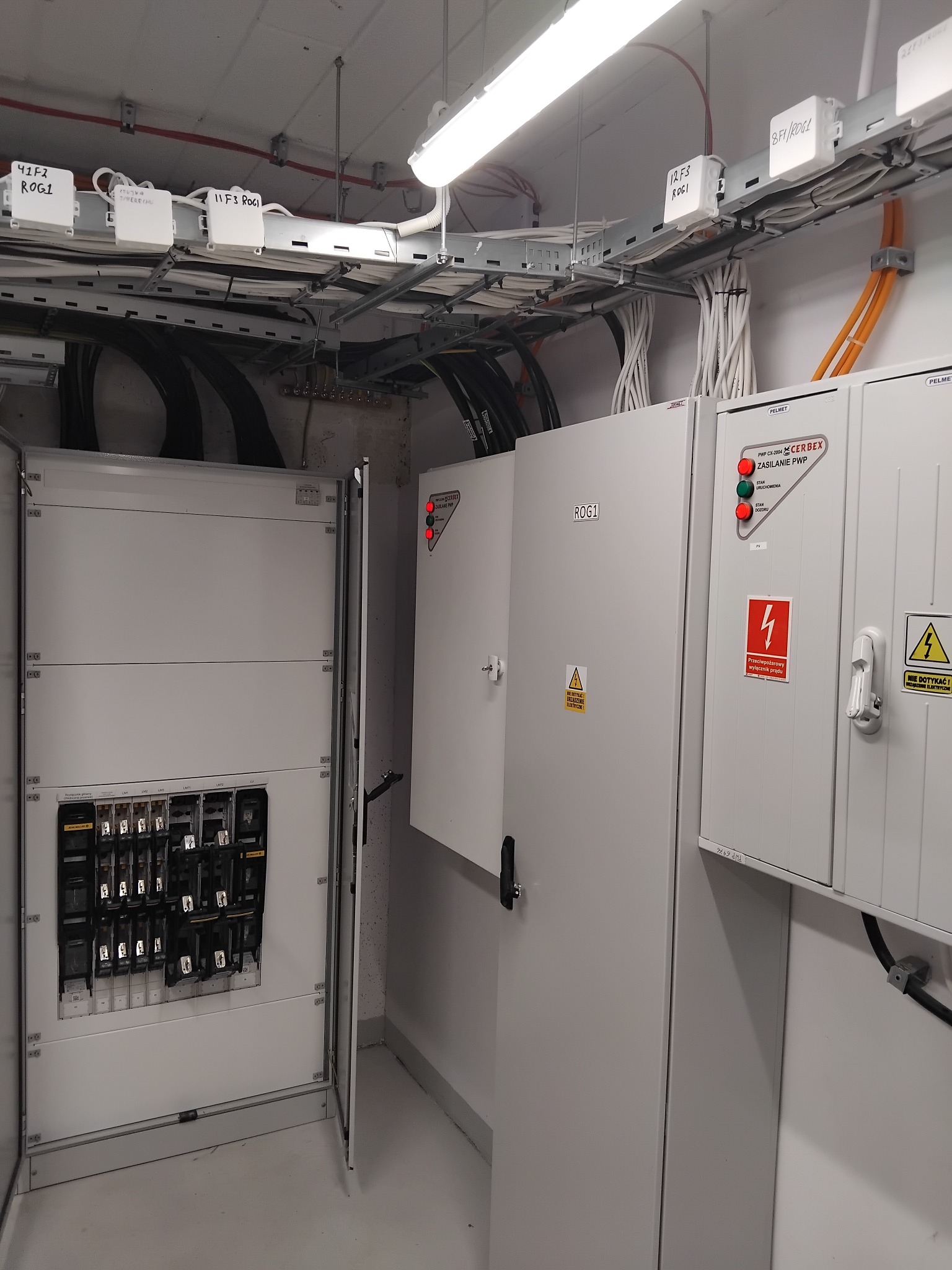 Rozdzielnia elektryczna z otwartymi drzwiami, widoczne zabezpieczenia i okablowanie. Tablice ostrzegawcze i oznaczenia. Instalacja w pomieszczeniu technicznym.