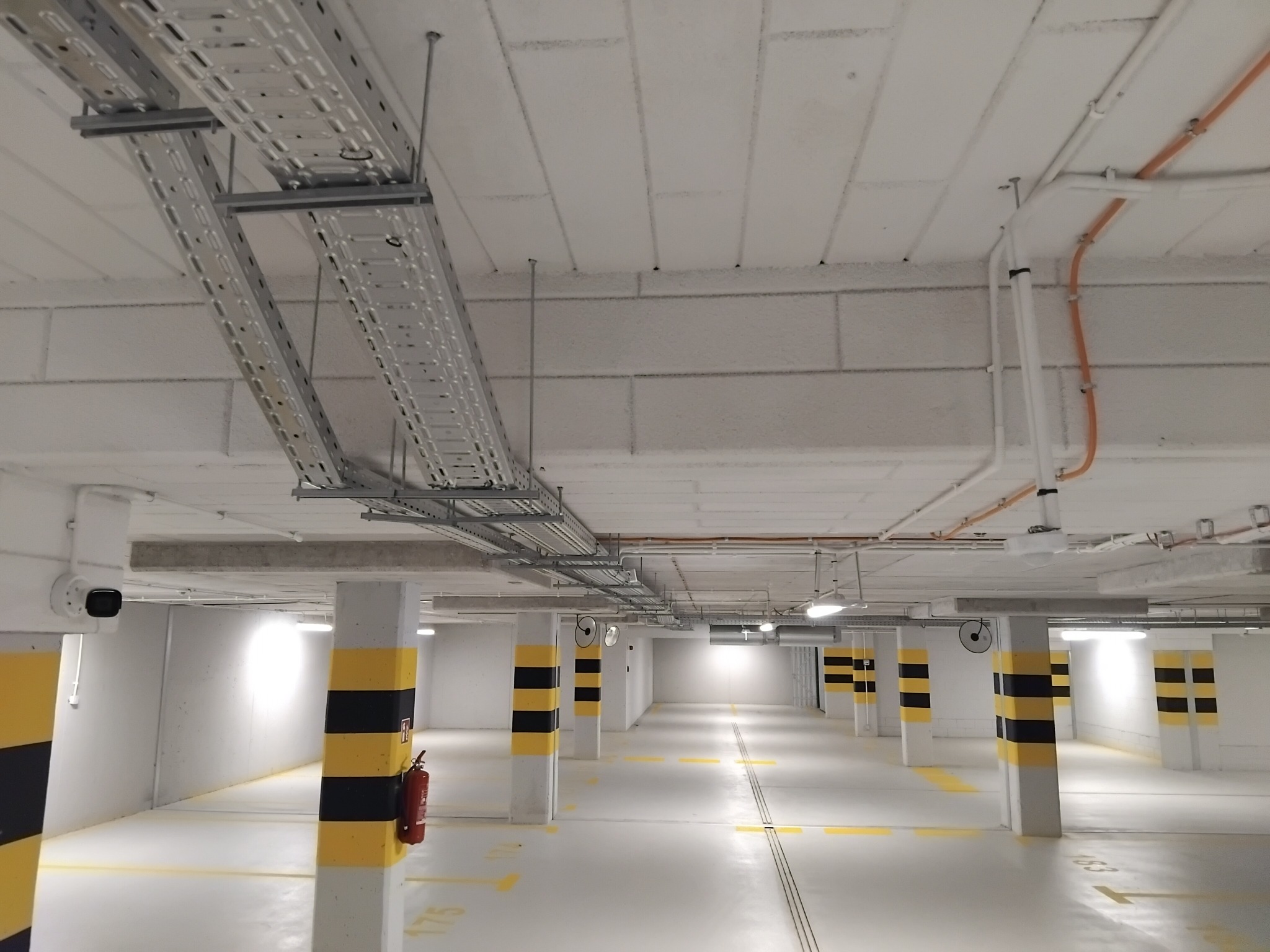 Kable i rury pod sufitem w garażu podziemnym. Widoczne metalowe korytka kablowe, rury w kolorze pomarańczowym i białym oraz słupy z pasami ostrzegawczymi.