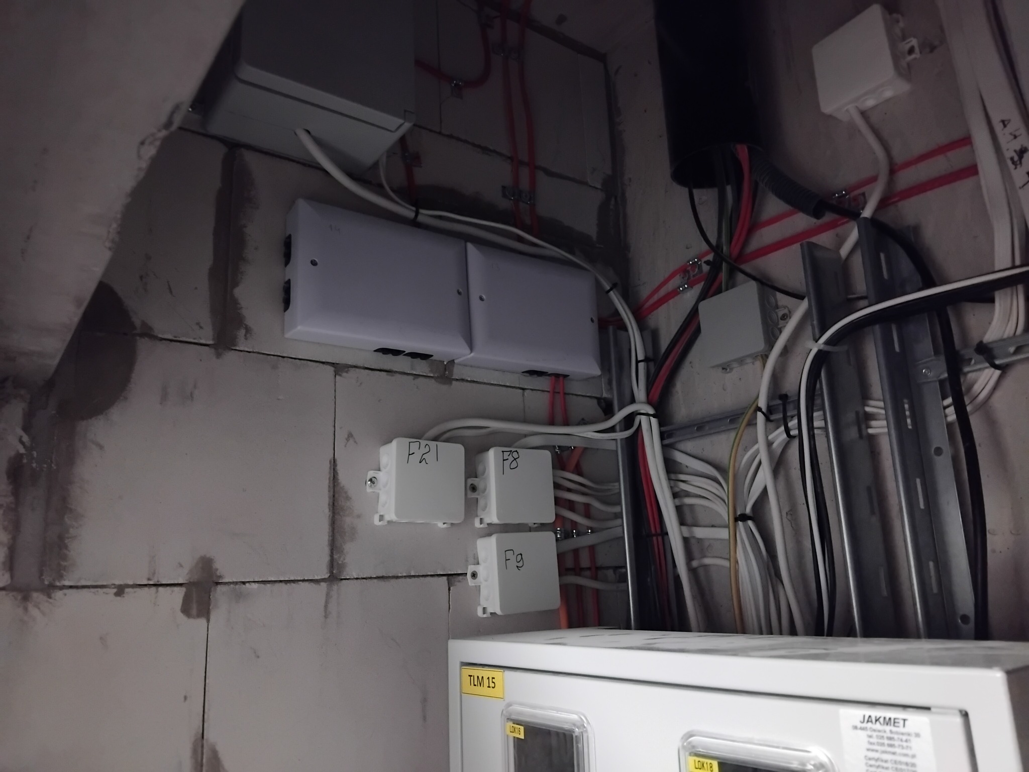 Rozdzielnia elektryczna z przewodami i puszkami na ścianie z betonu komórkowego. Widoczne oznaczenia na puszkach i etykieta informacyjna.