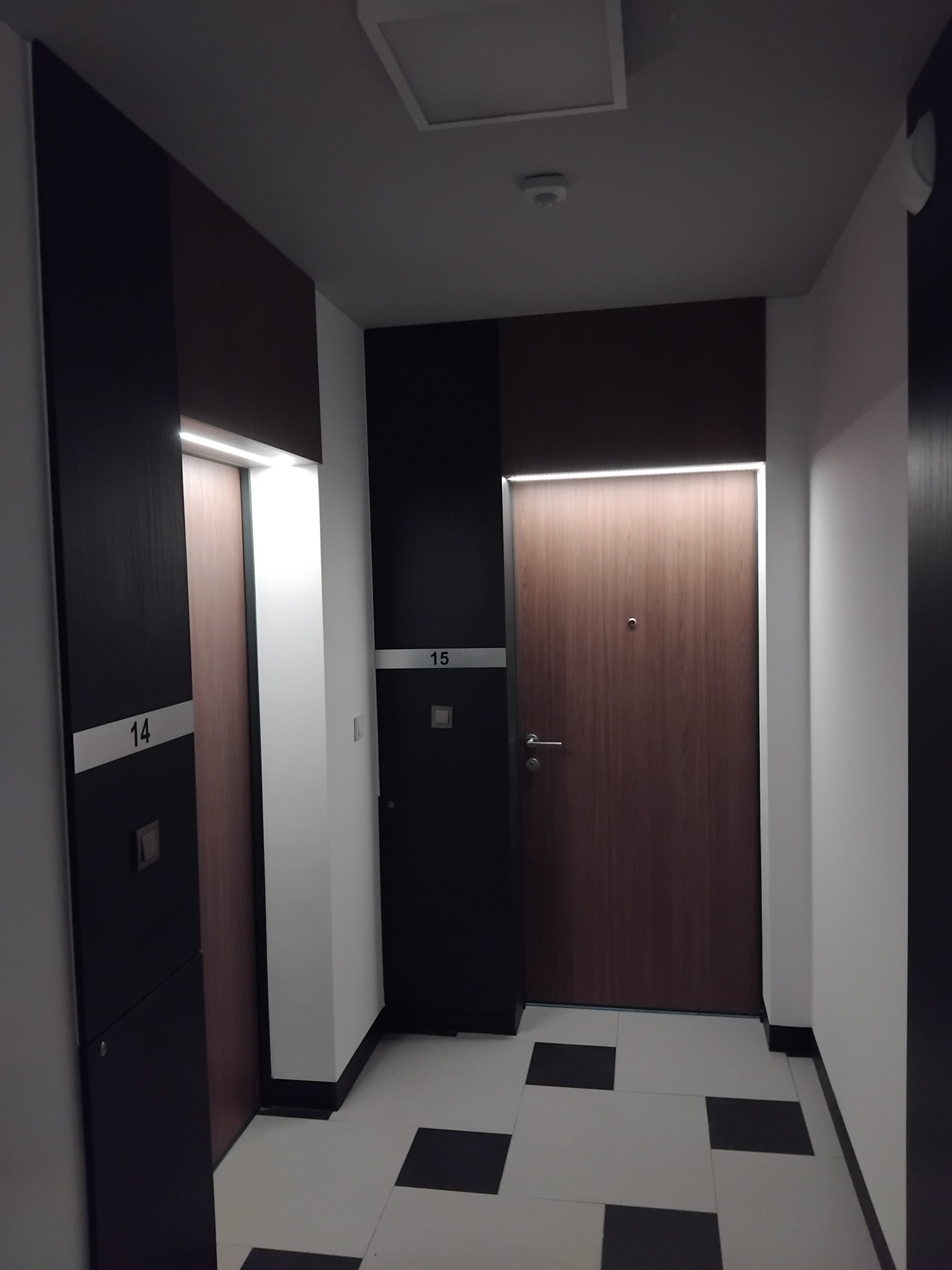 Korytarz w nowoczesnym stylu z drzwiami o numerach 14 i 15, oświetlonymi ledami. Czarno-biała podłoga, minimalistyczny design.