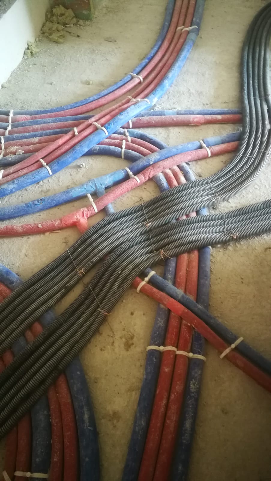 Instalacja podłogowa: rury grzewcze (czerwone i niebieskie) oraz peszle elektryczne ułożone na surowym betonie, zabezpieczone opaskami, gotowe do zalania posadzką.