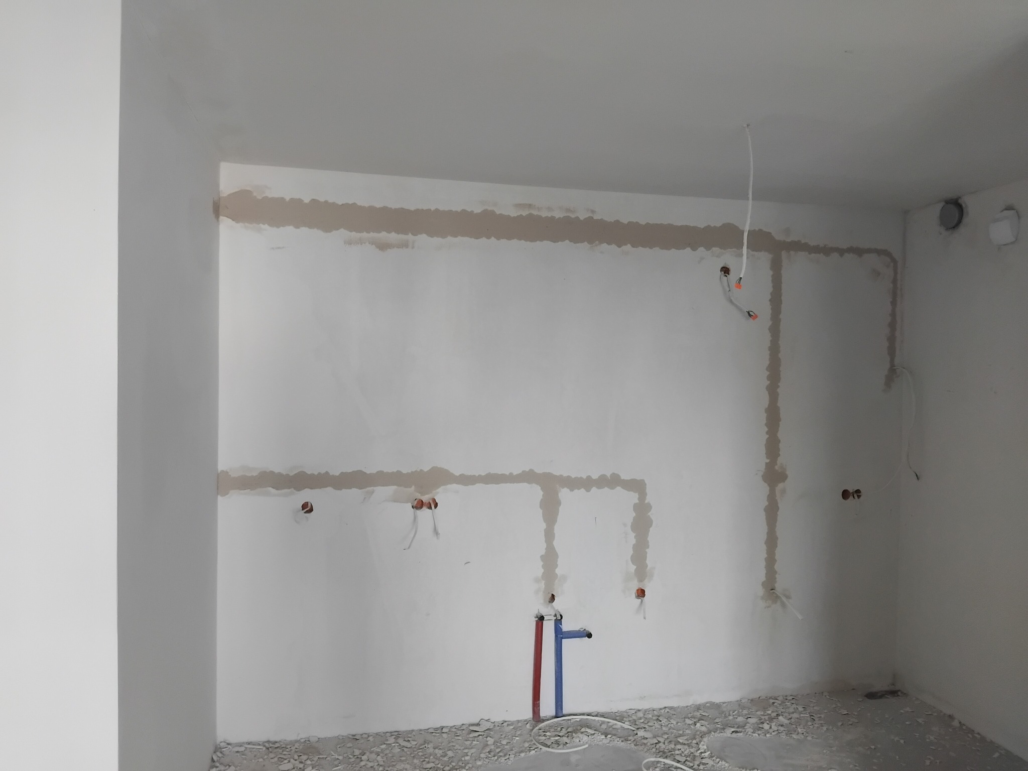 Ściana z instalacją elektryczną i hydrauliczną w trakcie prac wykończeniowych. Widoczne kable i rury wystające ze ściany, przygotowane do dalszych prac.