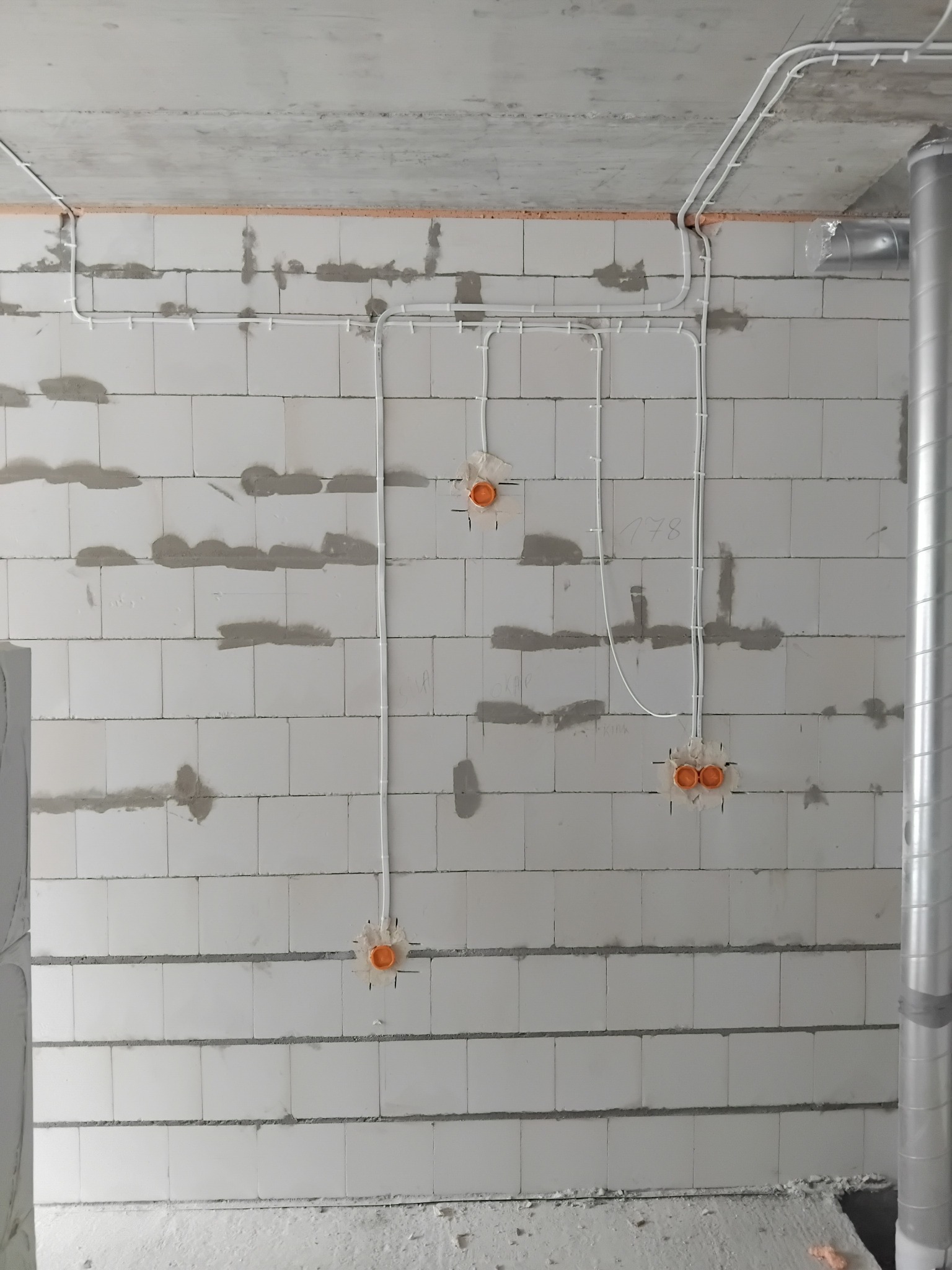 Surowa ściana z instalacją elektryczną: białe kable prowadzone po powierzchni, pomarańczowe puszki podtynkowe. Widoczny fragment wentylacji.