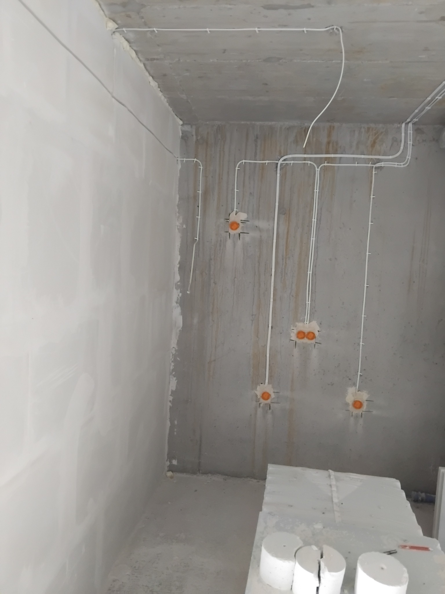 Surowy pokój z widoczną instalacją elektryczną: białe kable prowadzone po suficie i ścianach, pomarańczowe puszki elektryczne w ścianie. Widoczne materiały budowlane.