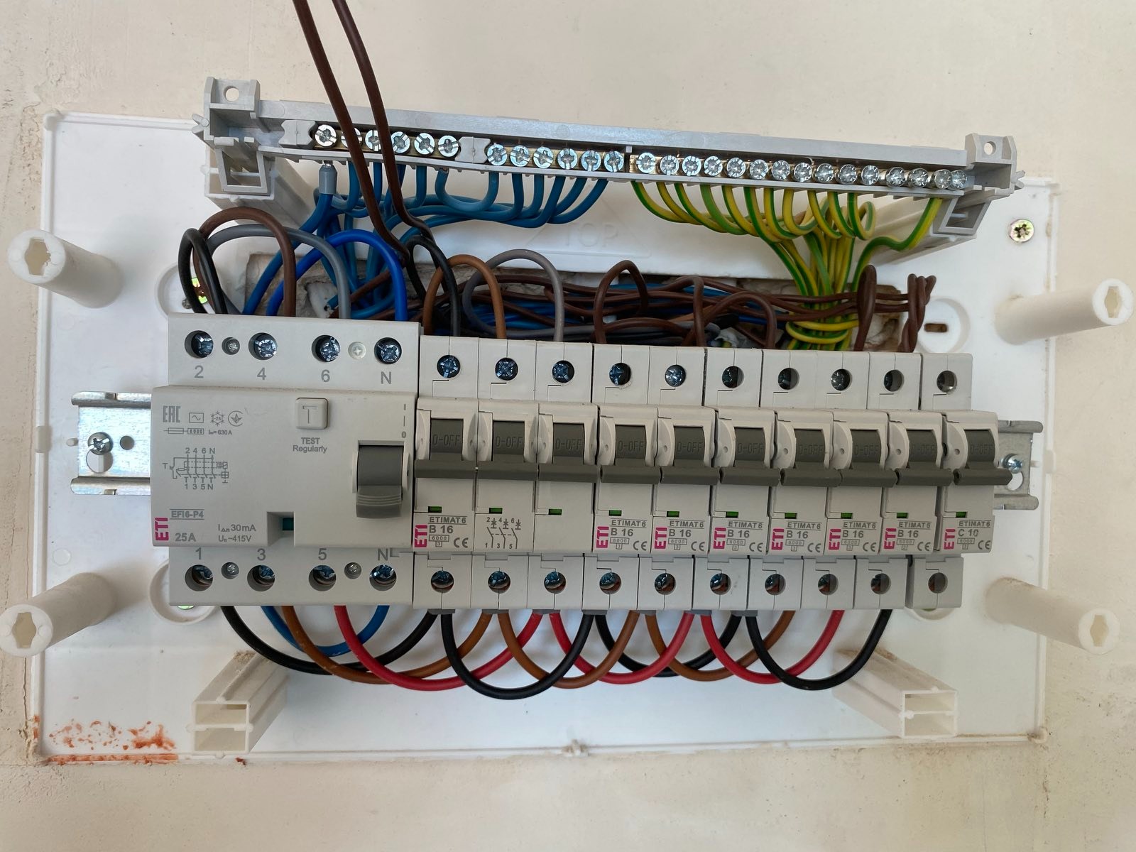 Otwarta skrzynka elektryczna z bezpiecznikami, widoczne różnokolorowe kable i listwa zaciskowa. Element instalacji wewnątrz budynku.