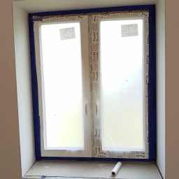 Grzegorz Kurek - Okno DAKO zabezpieczone niebieską taśmą malarską przed malowaniem. Na parapecie leży rolka folii ochronnej. Przygotowanie do prac wykończeniowych.