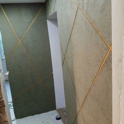 Grzegorz Kurek - Dekoracyjna ściana z szorstką fakturą i złotymi liniami tworzącymi geometryczny wzór. Widoczne przejście do innego pomieszczenia i fragment sufitu z oświetleniem LED.
