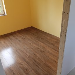 Grzegorz Kurek - Ukończone układanie paneli podłogowych w pokoju z żółtymi ścianami. Widoczny fragment okna i surowa ościeżnica drzwi z naniesionymi notatkami.