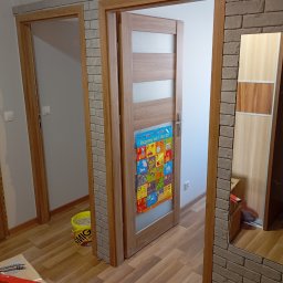 Grzegorz Kurek - Wykończone ościeżnice drzwiowe z dekoracyjną cegłą, widoczne narzędzia i wiadro z zaprawą. Układanie paneli podłogowych.